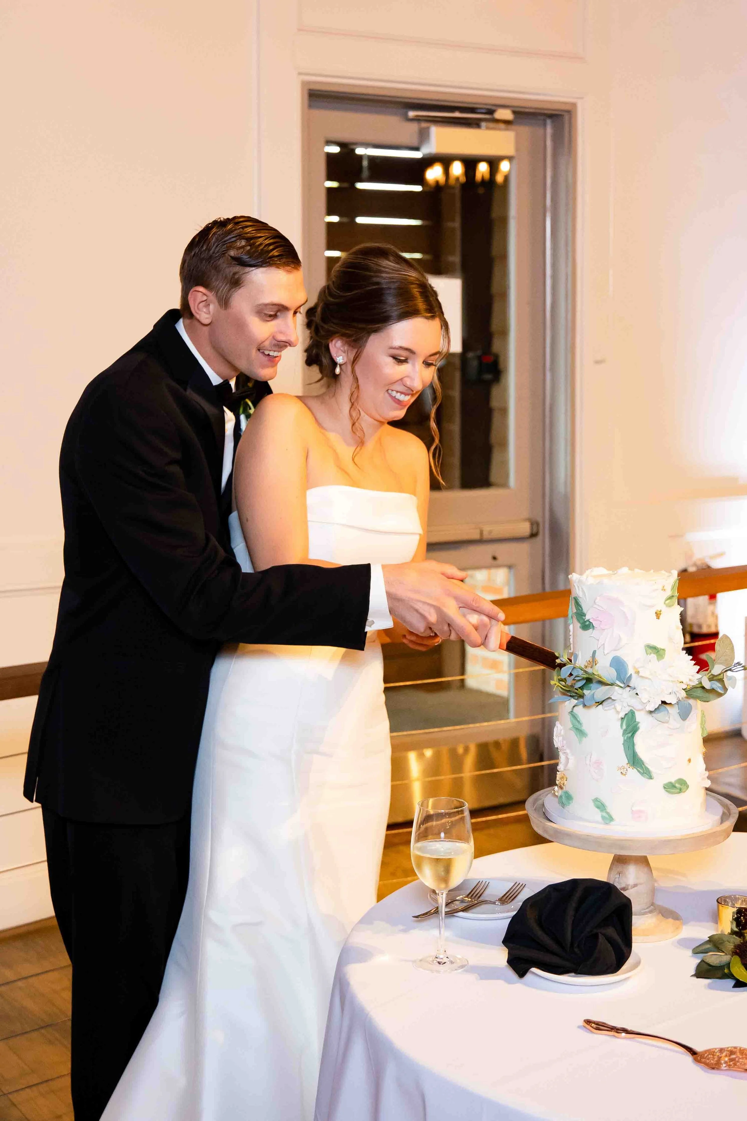 A newlywed couple in formal attire cutting a wedding cake together at their reception.
