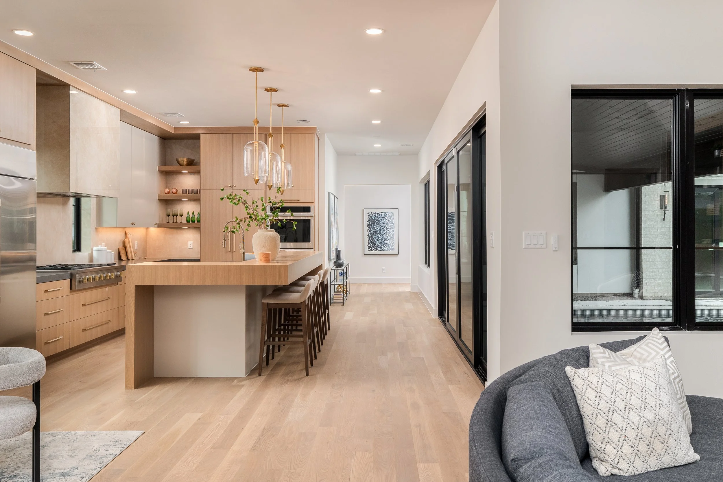 Modern kitchen with a light wood island, bar stools, and pendant lighting, open to a living space with a gray sofa and decorative pillows, black-framed sliding doors, and white walls.