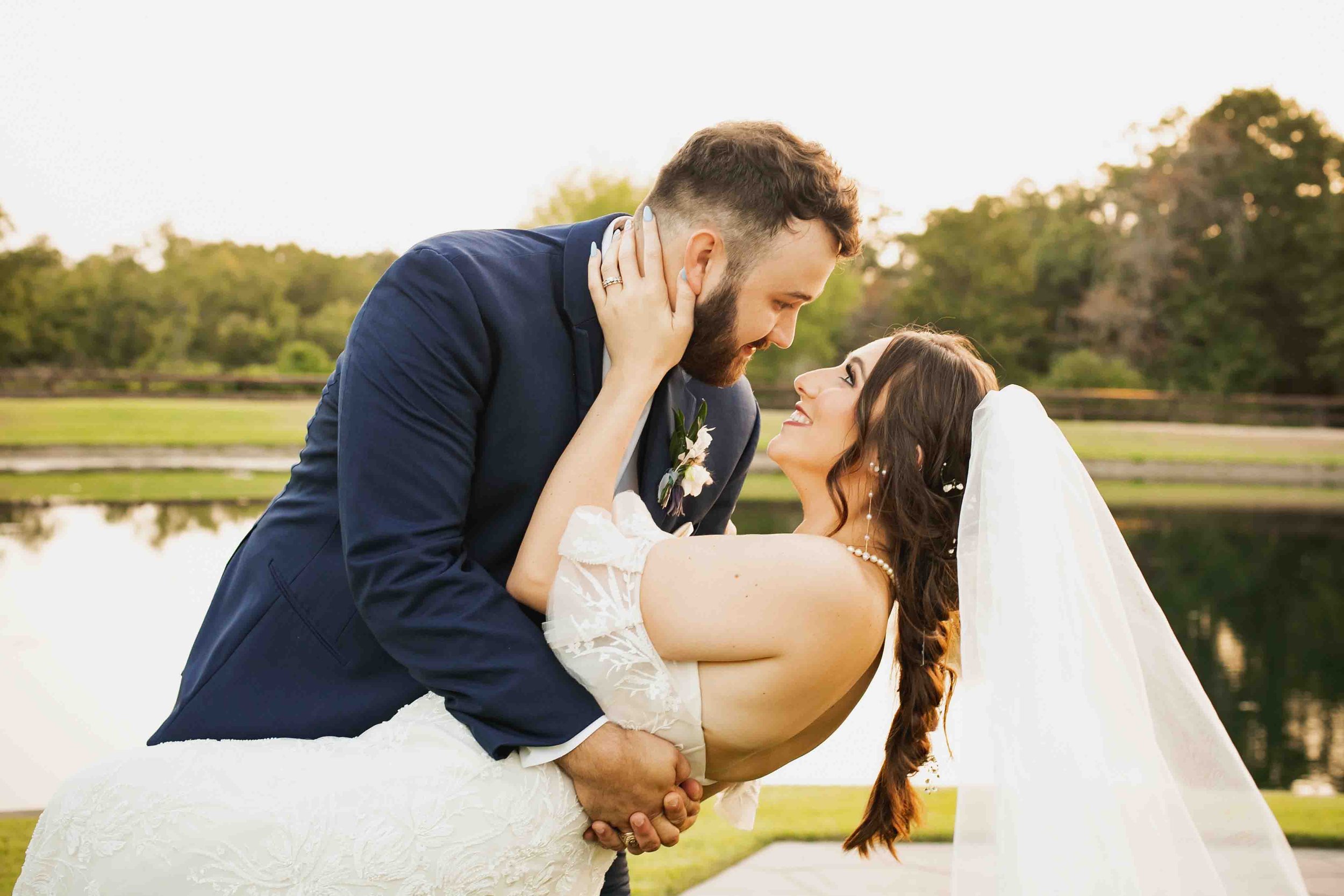 A couple in wedding attire celebrating outdoors by a lake, with the groom dipping the bride and holding her waist, as they gaze into each other's eyes.