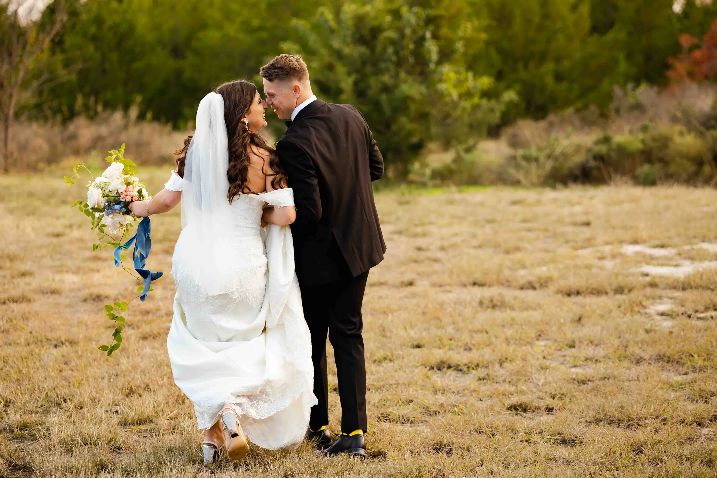 A bride and groom dancing outdoors on a grassy field with trees in the background, smiling and close to each other.