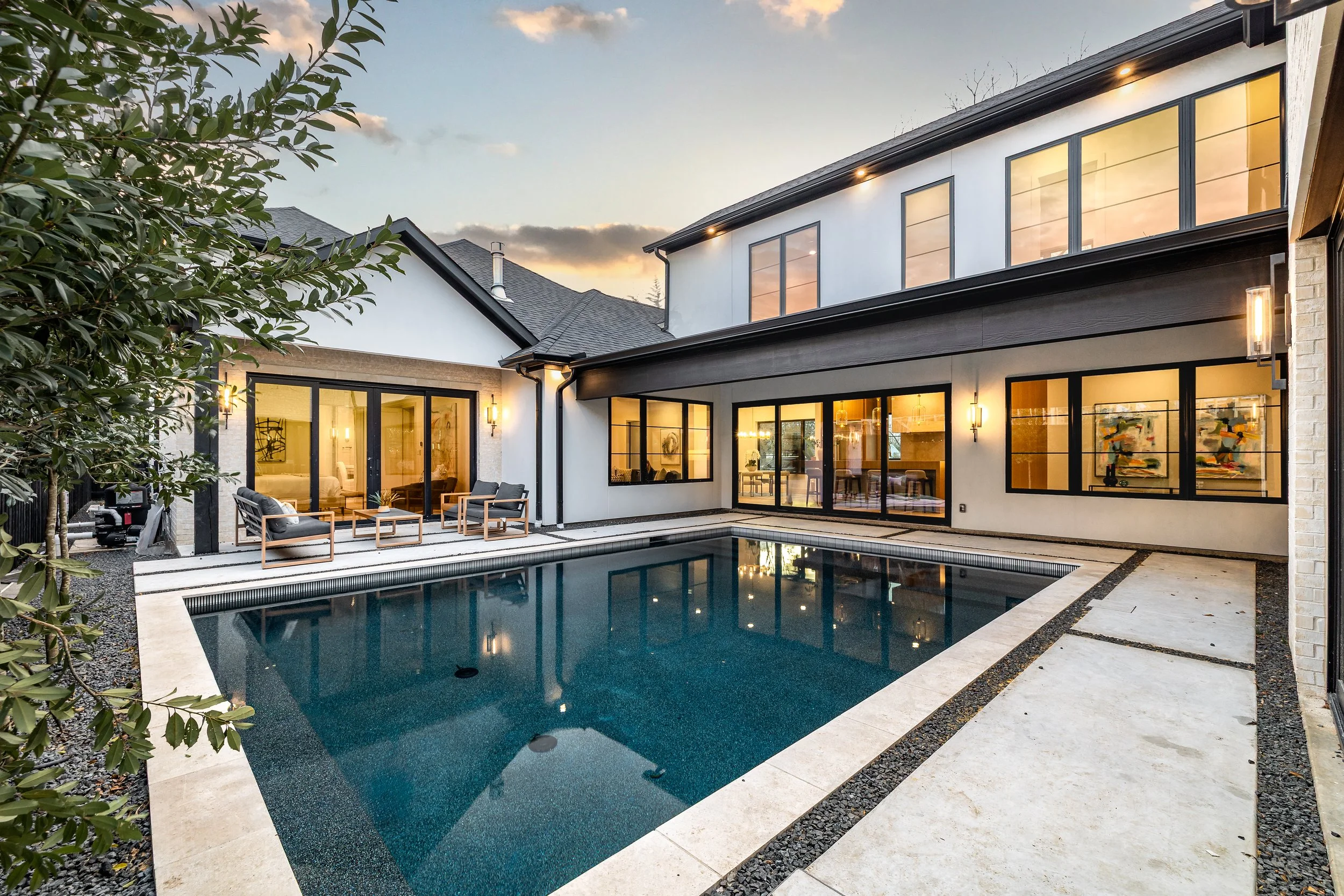 Modern house with large glass windows and a backyard swimming pool at sunset, outdoor sitting area with chairs and tables.