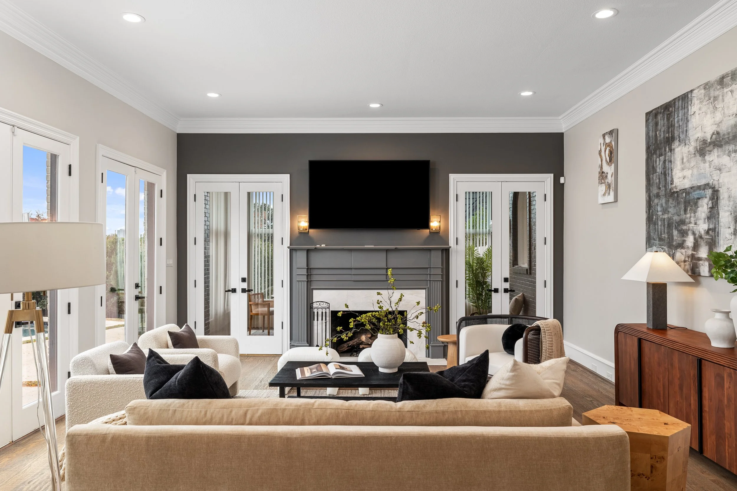 Living room with beige sofa, black pillows, armchair, white vases, coffee table, fireplace, TV, glass doors, artwork, and wooden sideboard.