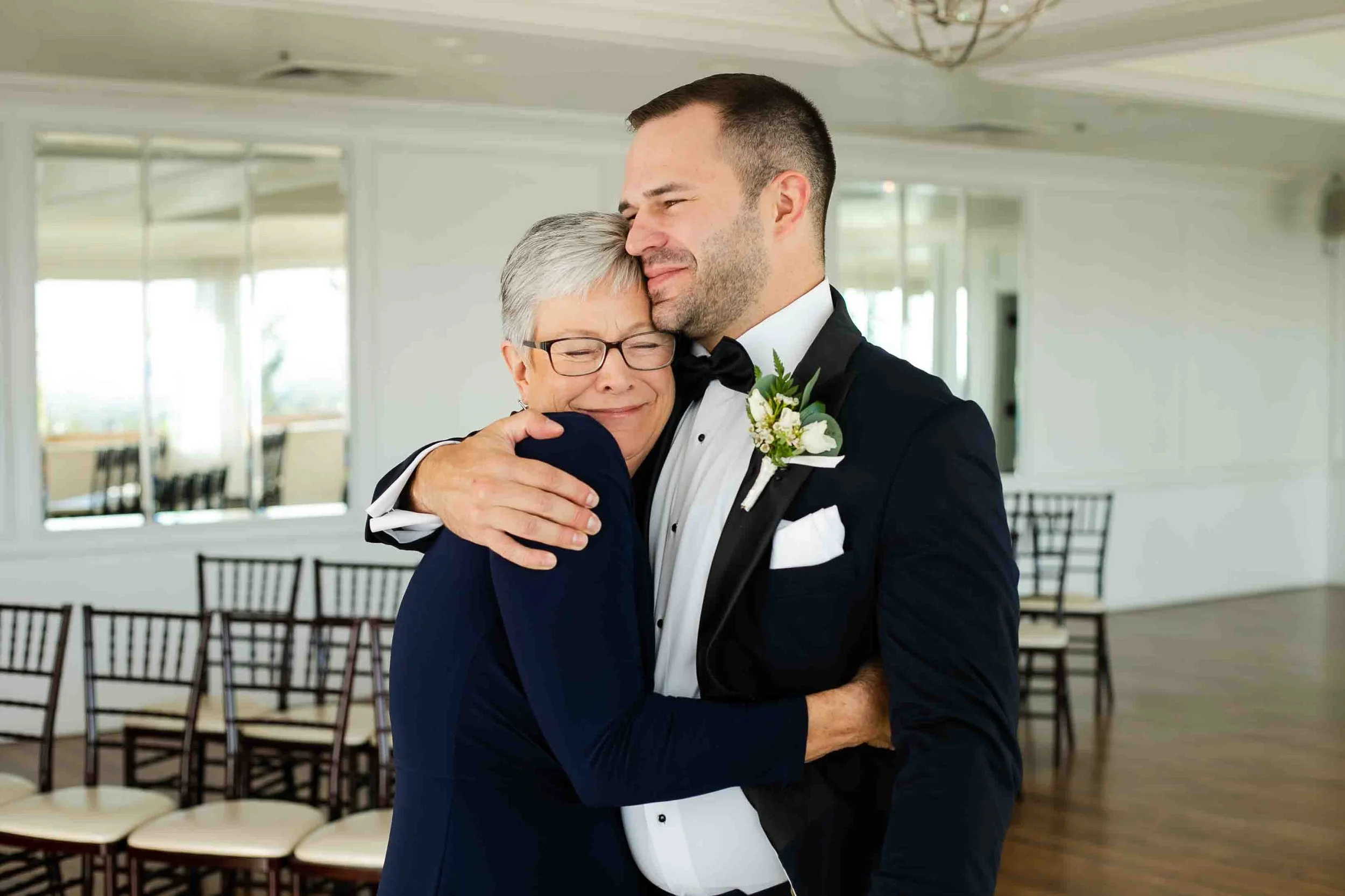 A young man in a black tuxedo with a boutonnière hugs an older woman in a navy dress, both smiling with eyes closed, in a bright room with chairs and mirrors.