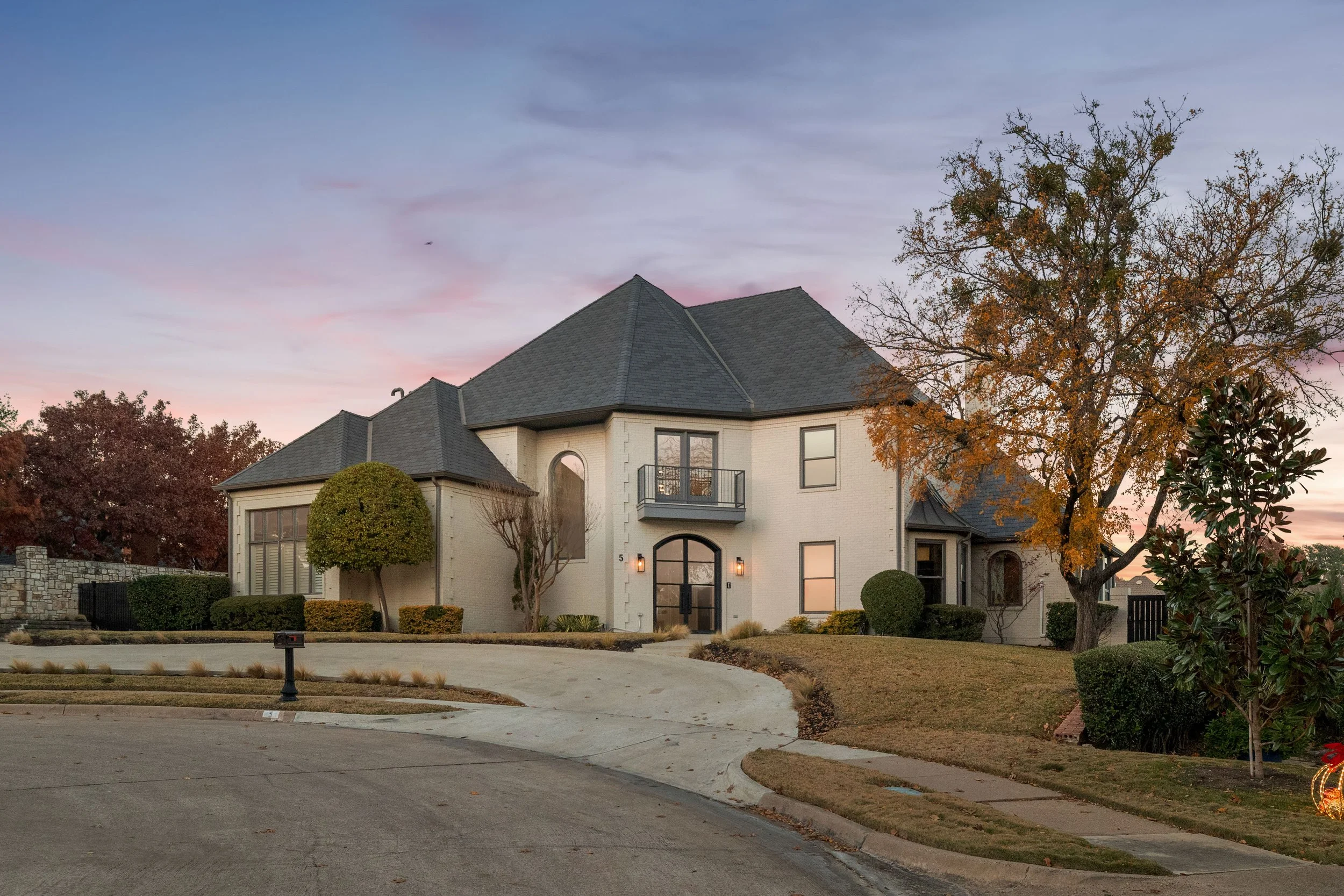 Two-story modern house with white exterior, black roof, and front door with lights; landscaped yard with trees, bushes, and a curved driveway at sunset.