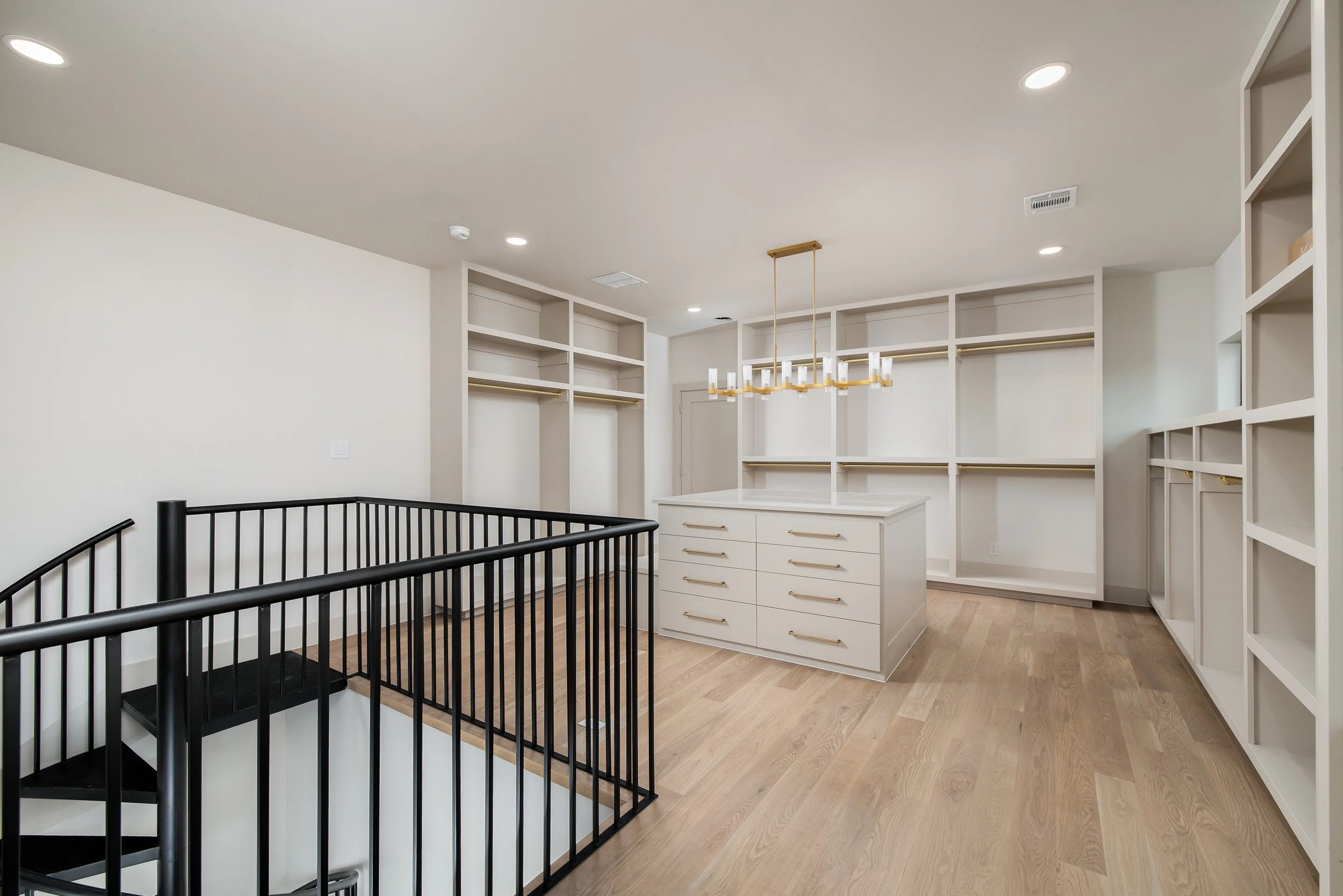 Empty walk-in closet with built-in white shelving units, a white island with drawers, and a modern chandelier, with beige wood flooring and recessed ceiling lights.