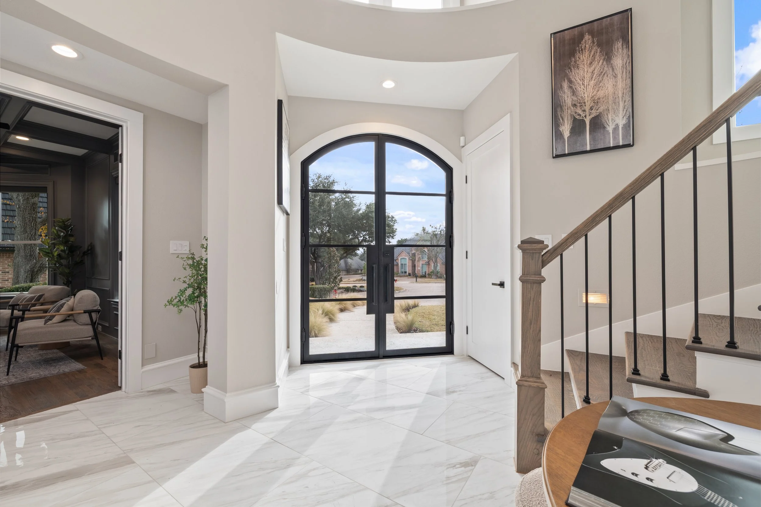 Modern home entryway with a black framed glass door, marble floor, staircase with wooden handrail, and minimalist wall art.