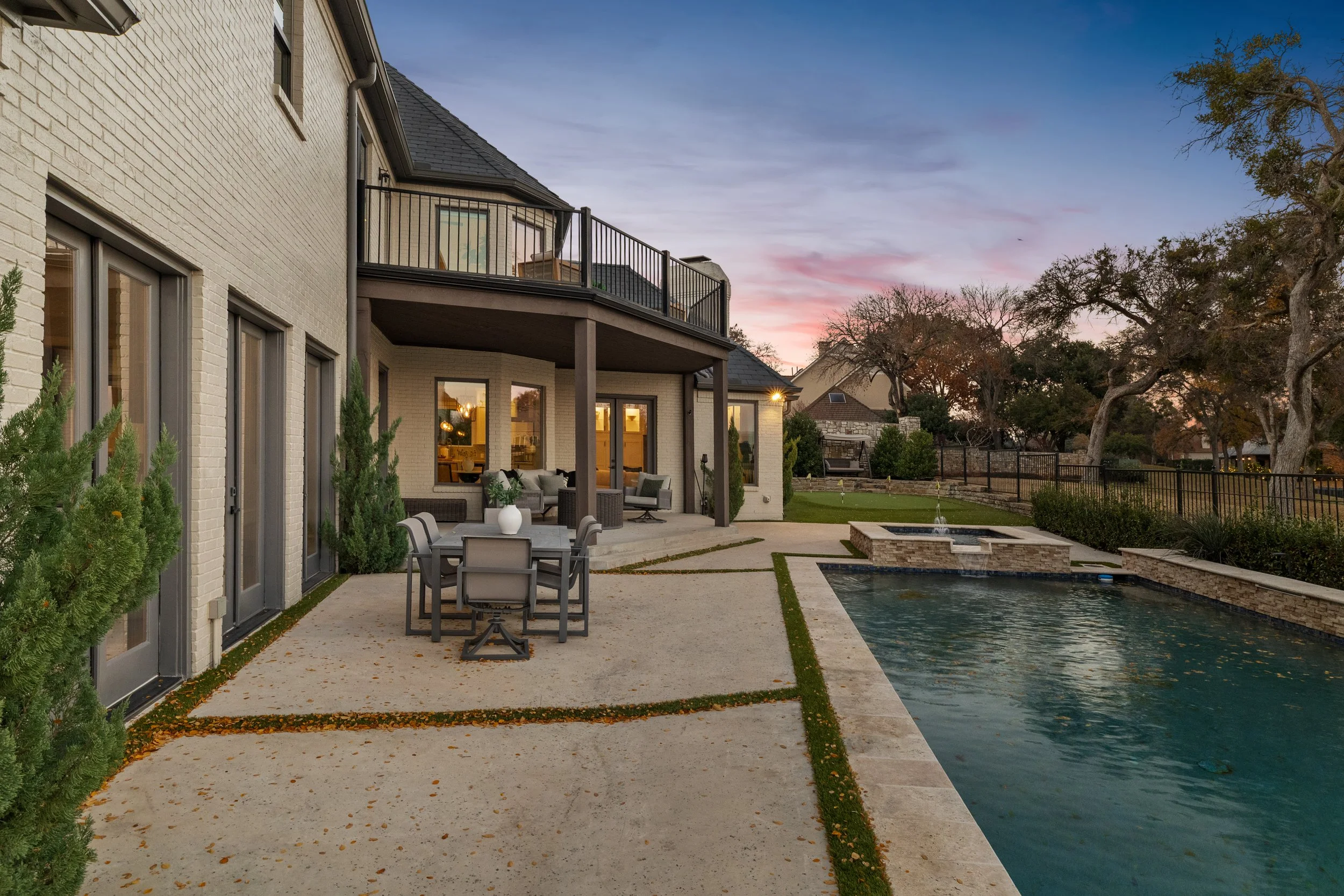 Backyard with pool, patio furniture, and house with balcony at dusk, trees, and fence.