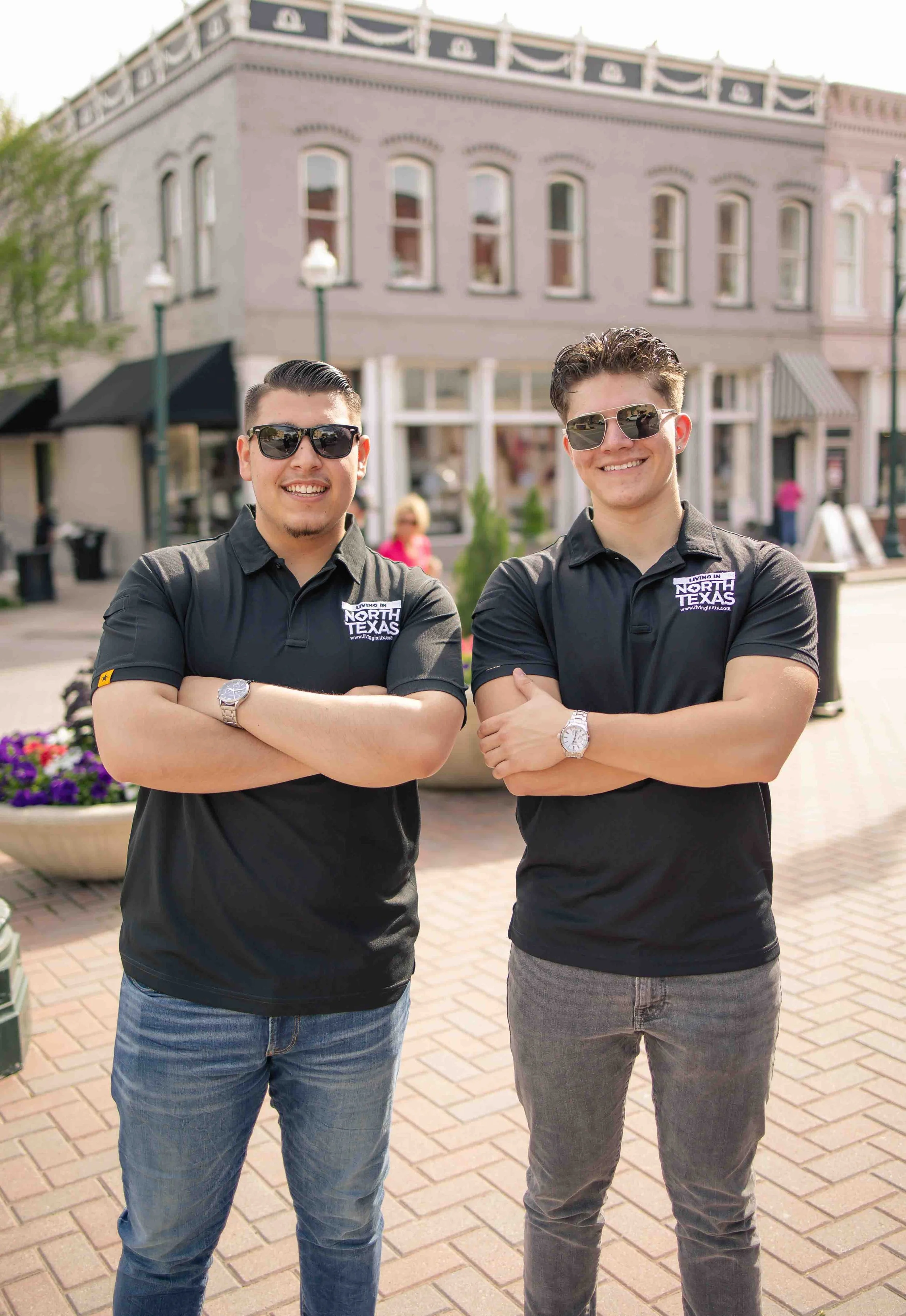 Two young men standing on a brick sidewalk in an outdoor shopping area, wearing black polo shirts with 'Living in North Texas' logos, both smiling and wearing sunglasses, with one in blue jeans and the other in gray jeans.