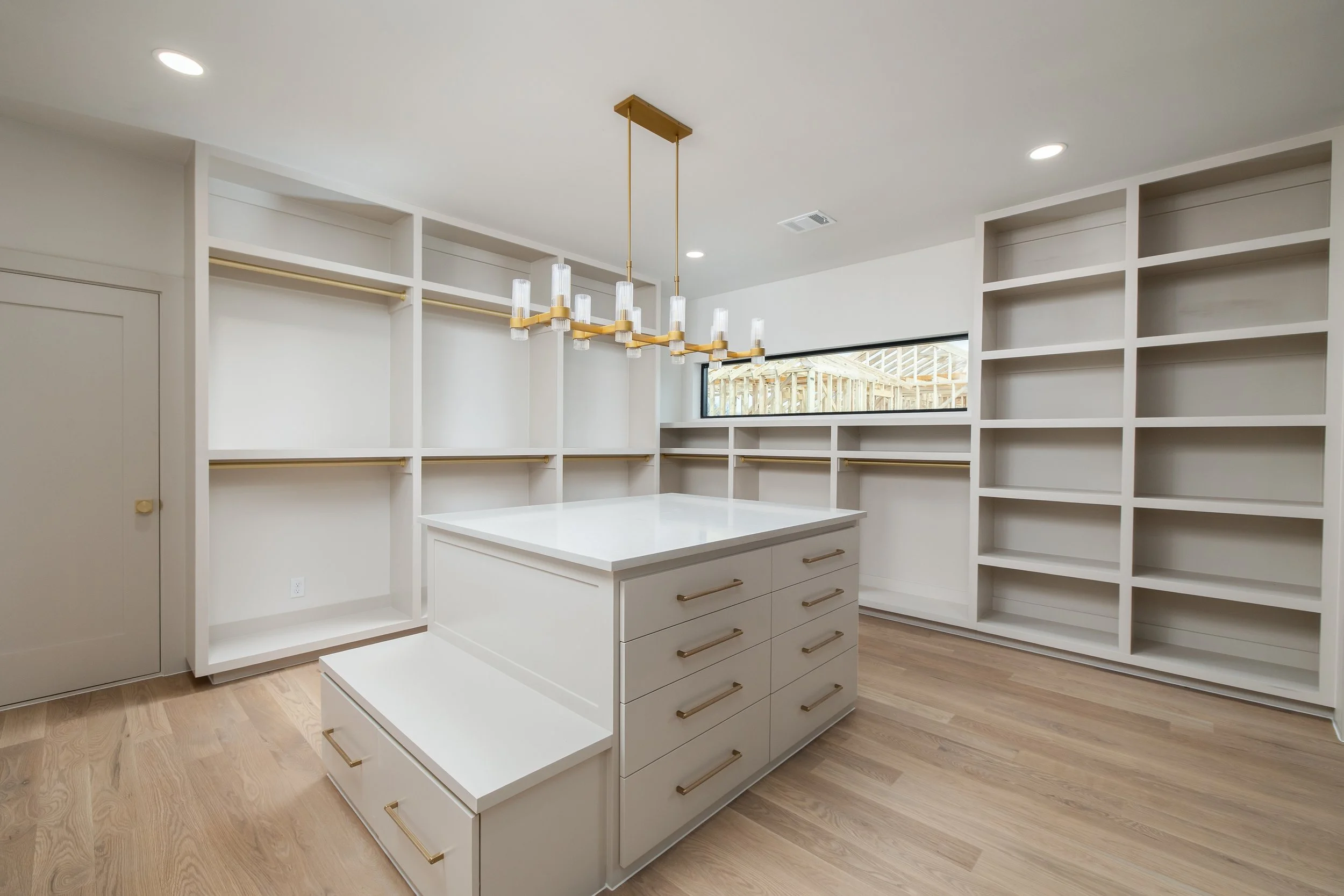 Empty walk-in closet with white built-in shelves, drawers, and a central island, hardwood floors, and a modern chandelier, with a long horizontal window.