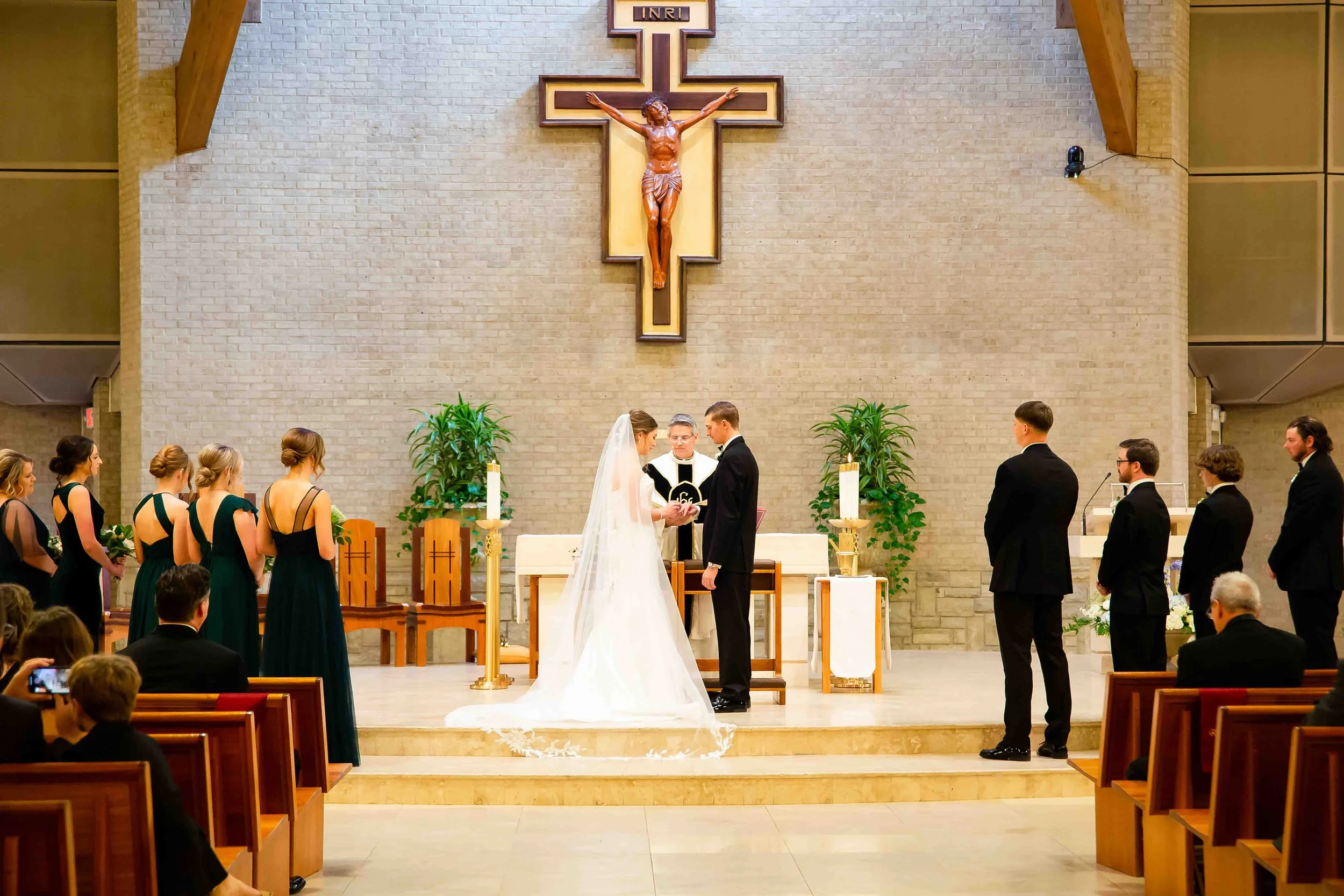 A wedding ceremony taking place inside a church with a crucifix on the wall. The bride and groom stand before the officiant, with bridesmaids and groomsmen on either side.