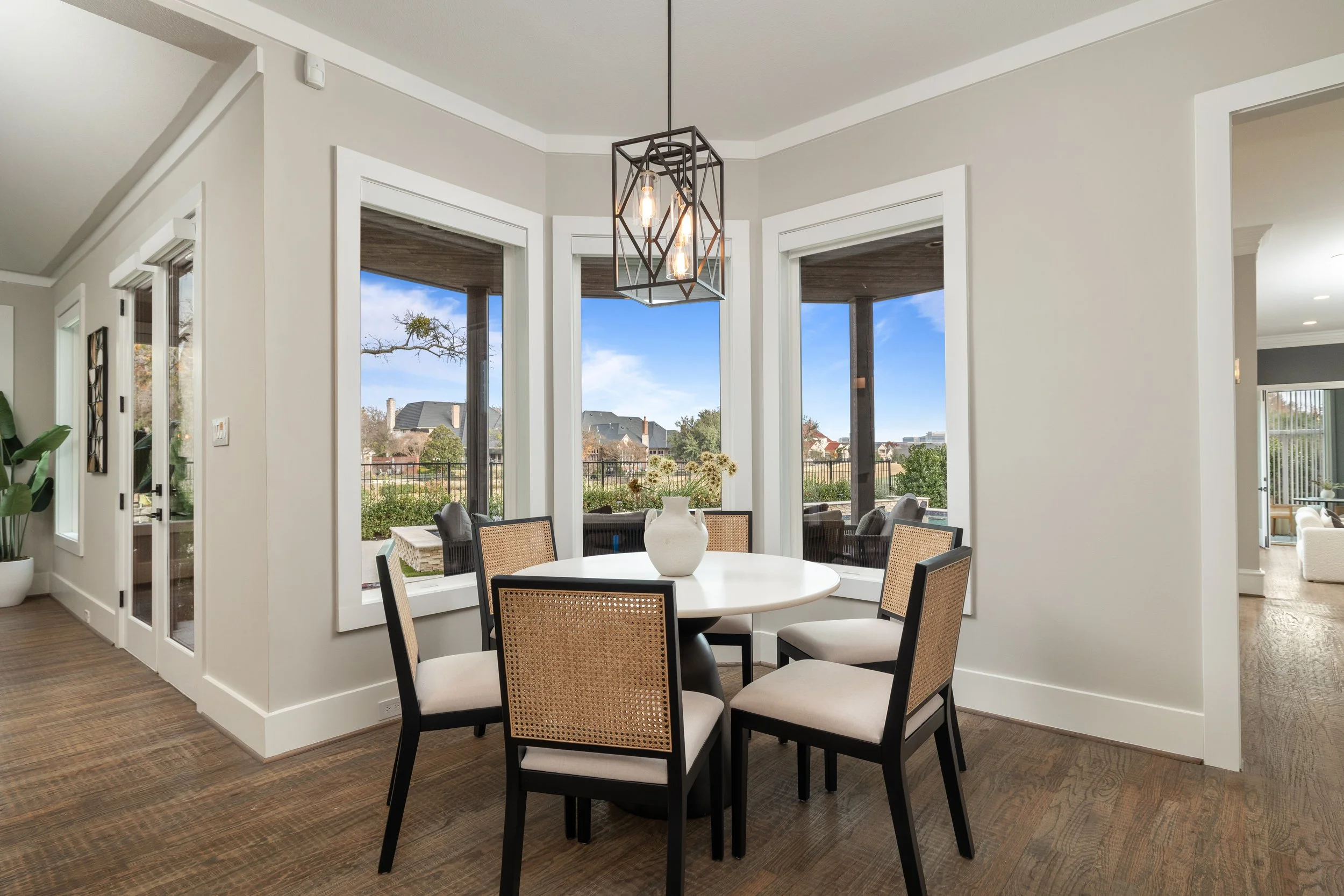 Dining room with round white table, six chairs with woven backs, large windows overlooking outdoor patio and yard, modern geometric pendant light hanging above table, hardwood floors, white walls.