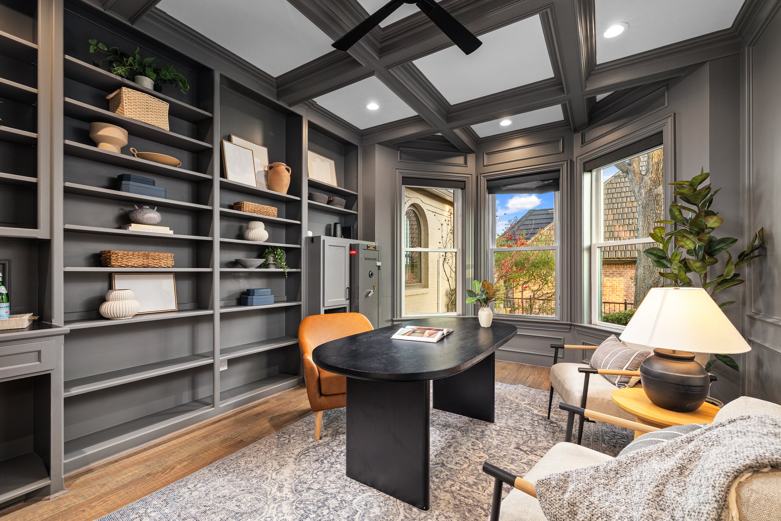 A home office with gray built-in bookshelves, a black desk, an orange armchair, a beige armchair, and large windows showing a tree and neighboring house with a shingled roof.