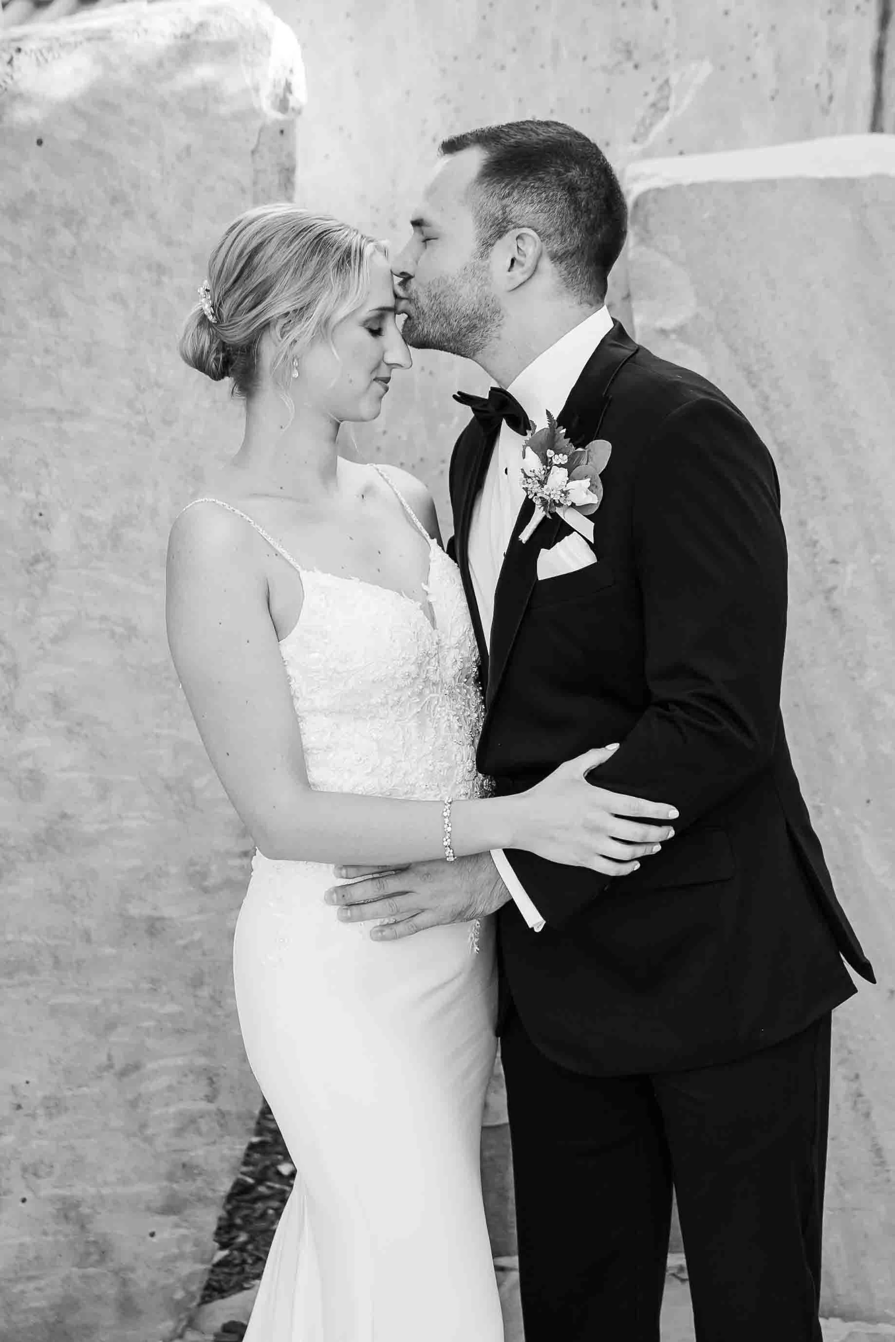 A bride and groom share a tender moment on their wedding day, with their foreheads pressed together and eyes closed, standing against a textured stone wall.