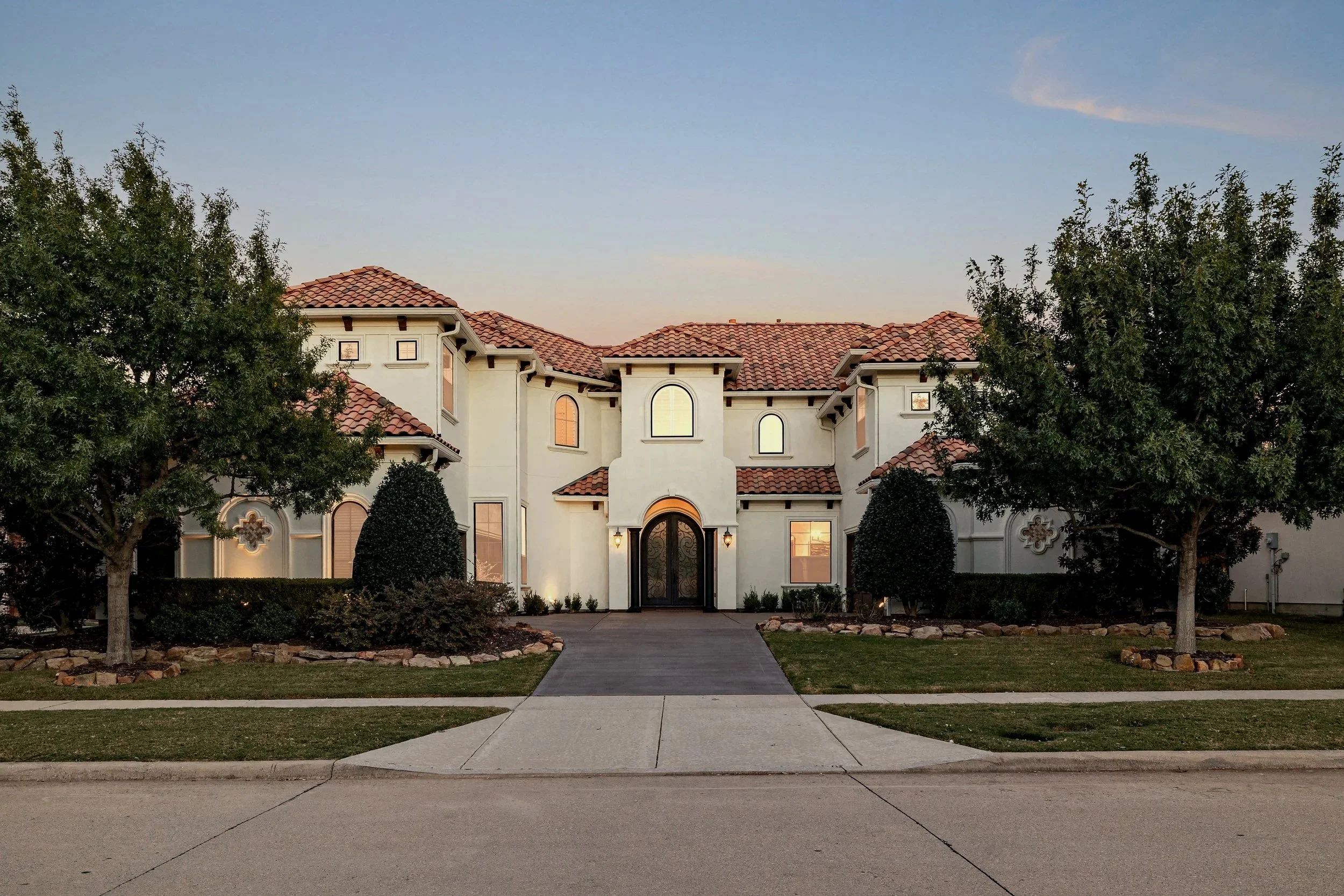 A large, white two-story house with a red tile roof, arched windows, and a decorative front door, surrounded by well-maintained landscaping and trees.
