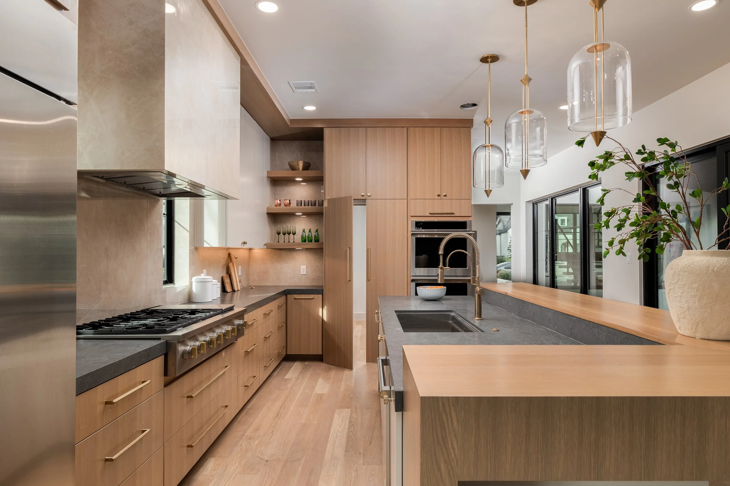 Modern kitchen with light wood cabinetry, a large dark countertop island with a sink and gold faucet, pendant lights hanging above, and sliding glass doors leading outside.