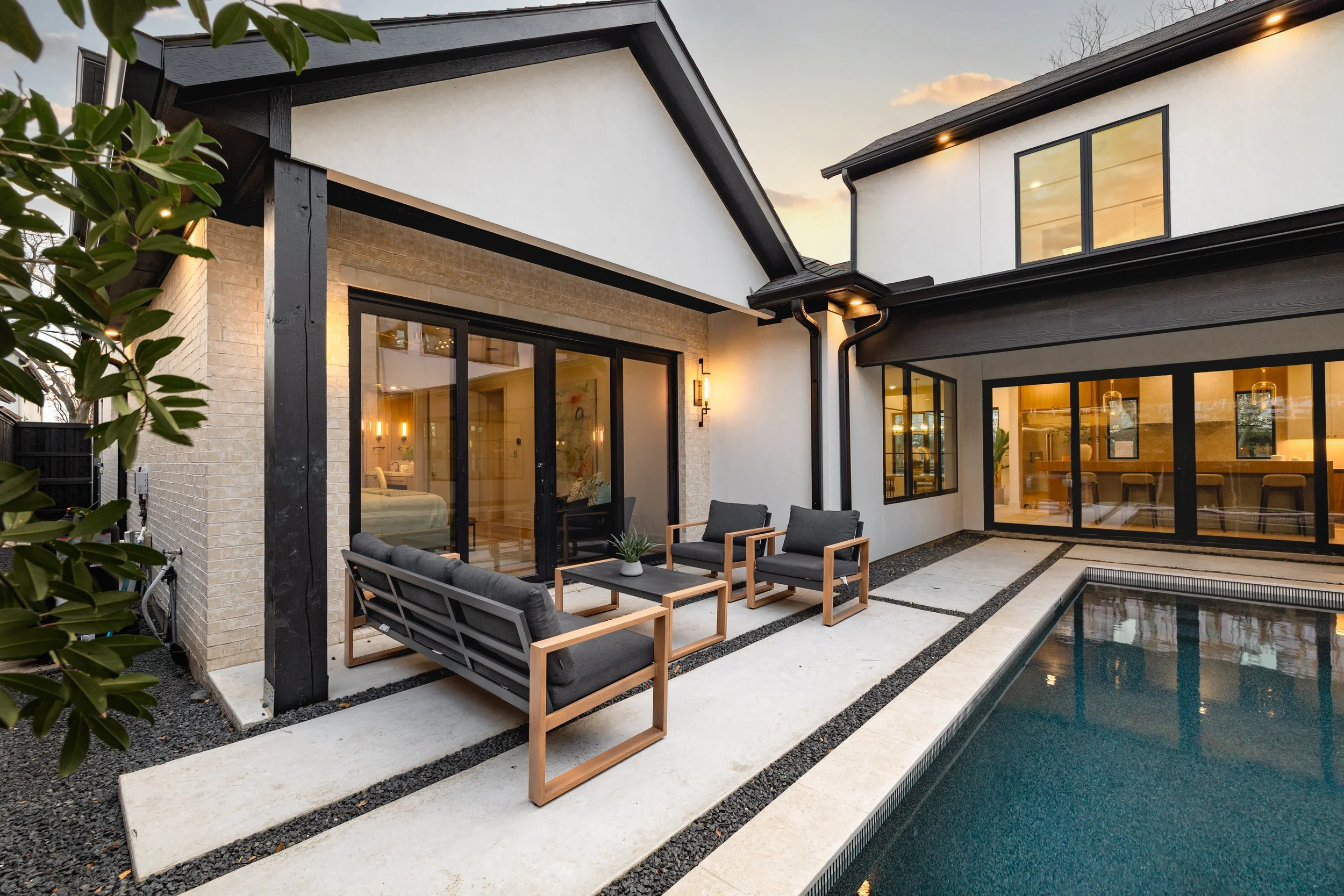 Modern house backyard with a swimming pool, outdoor seating area with black cushions, and large glass sliding doors and windows reflecting indoor lighting at sunset.