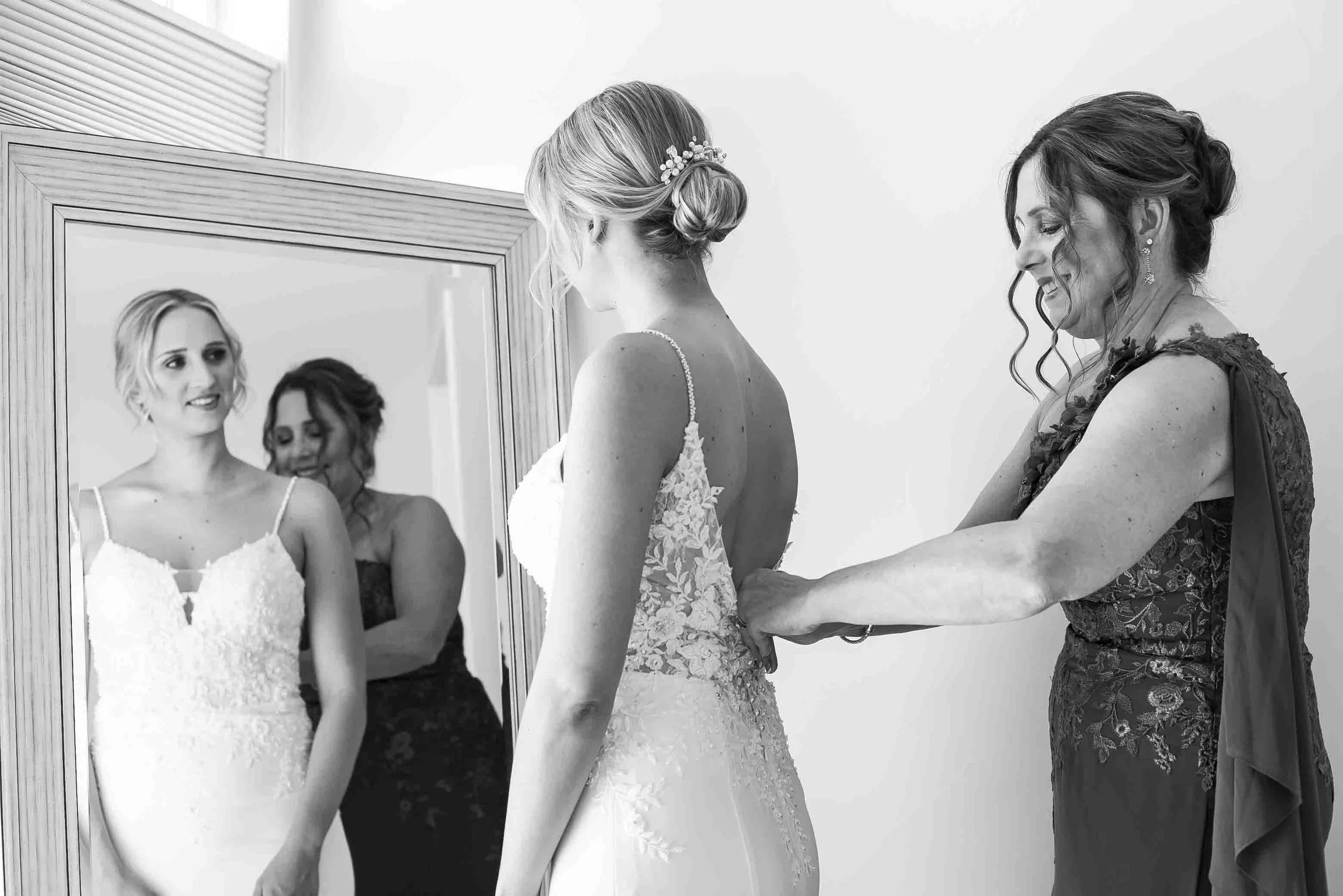 A bride getting ready with her mother or a helper adjusting her wedding dress in front of a mirror.