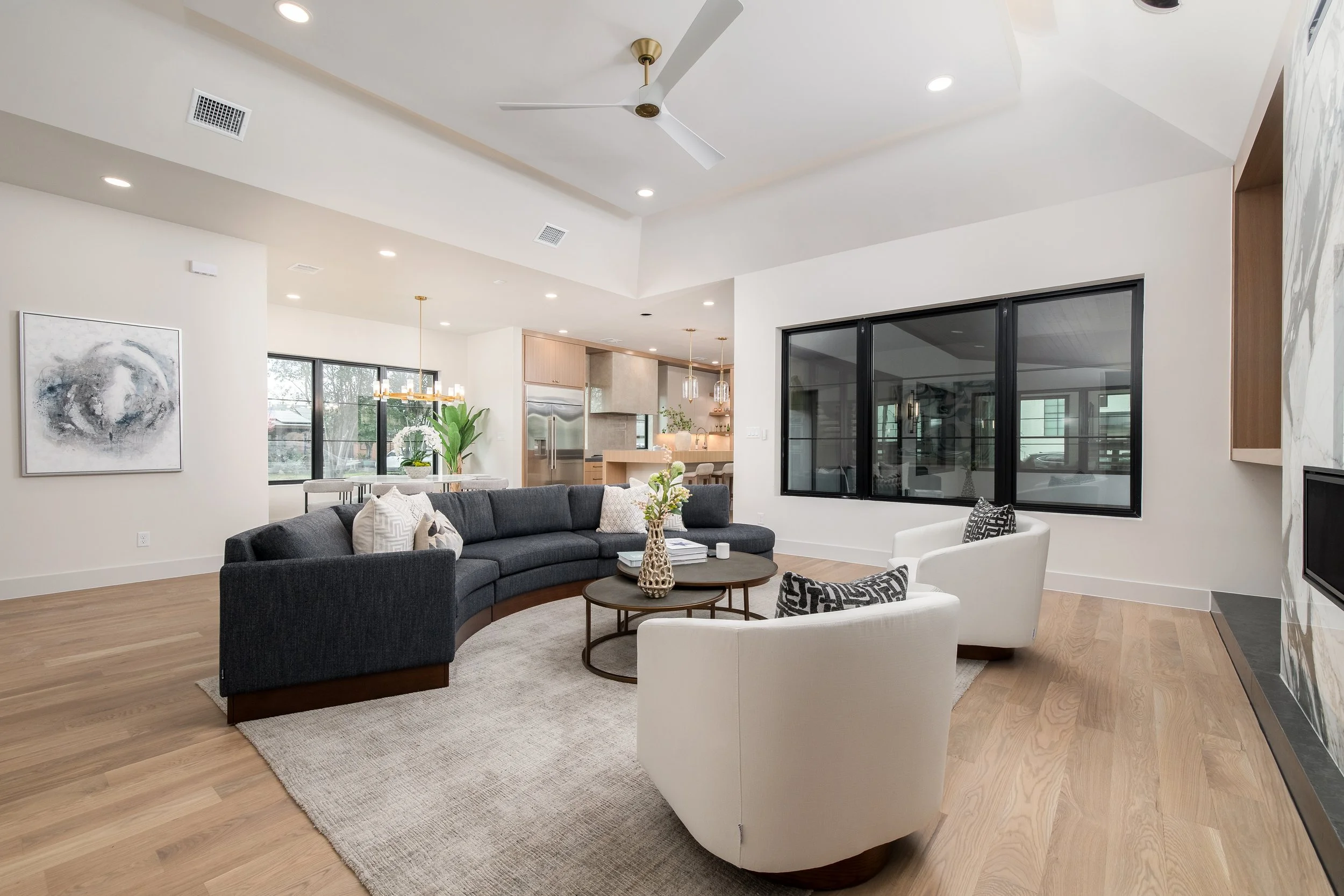 Modern living room with a curved dark gray sofa, two white armchairs, a round coffee table, and large windows