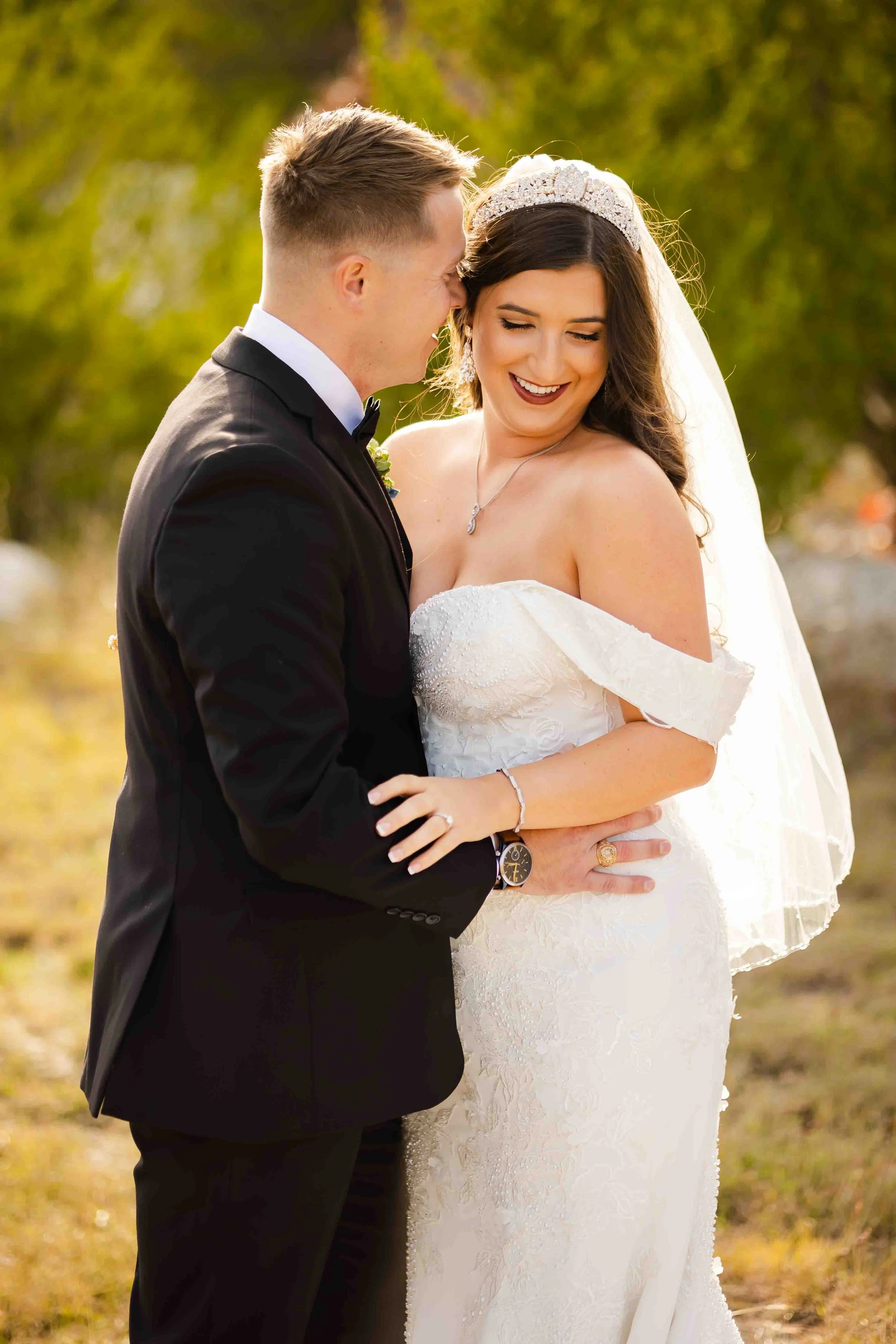 A bride and groom sharing an intimate moment outdoors, with the groom in a black tuxedo and the bride in a white wedding gown and veil, surrounded by greenery.