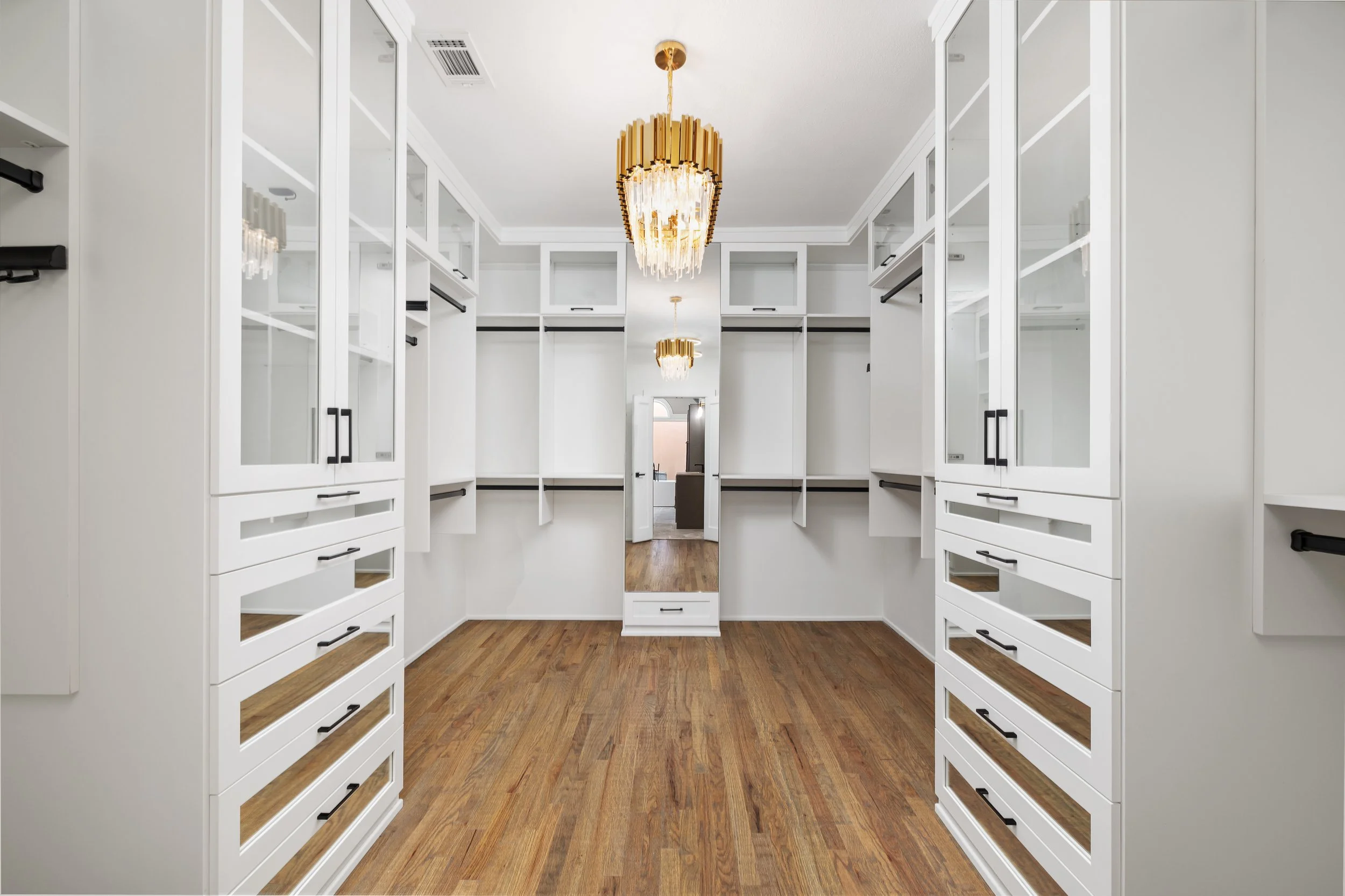 A spacious walk-in closet with white shelves and cabinets, hardwood floors, a mirror in the center, and two gold chandeliers hanging from the ceiling.