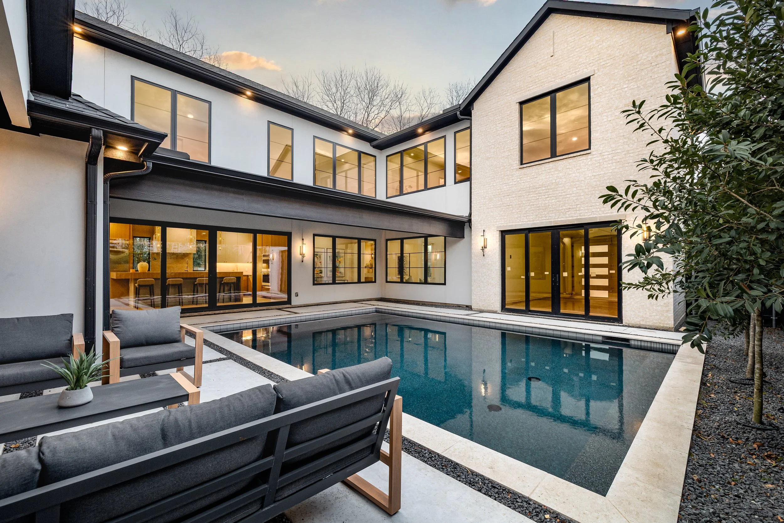 Modern two-story house with large windows, a backyard swimming pool, and outdoor seating area with cushioned chairs and a small plant, during evening hours.