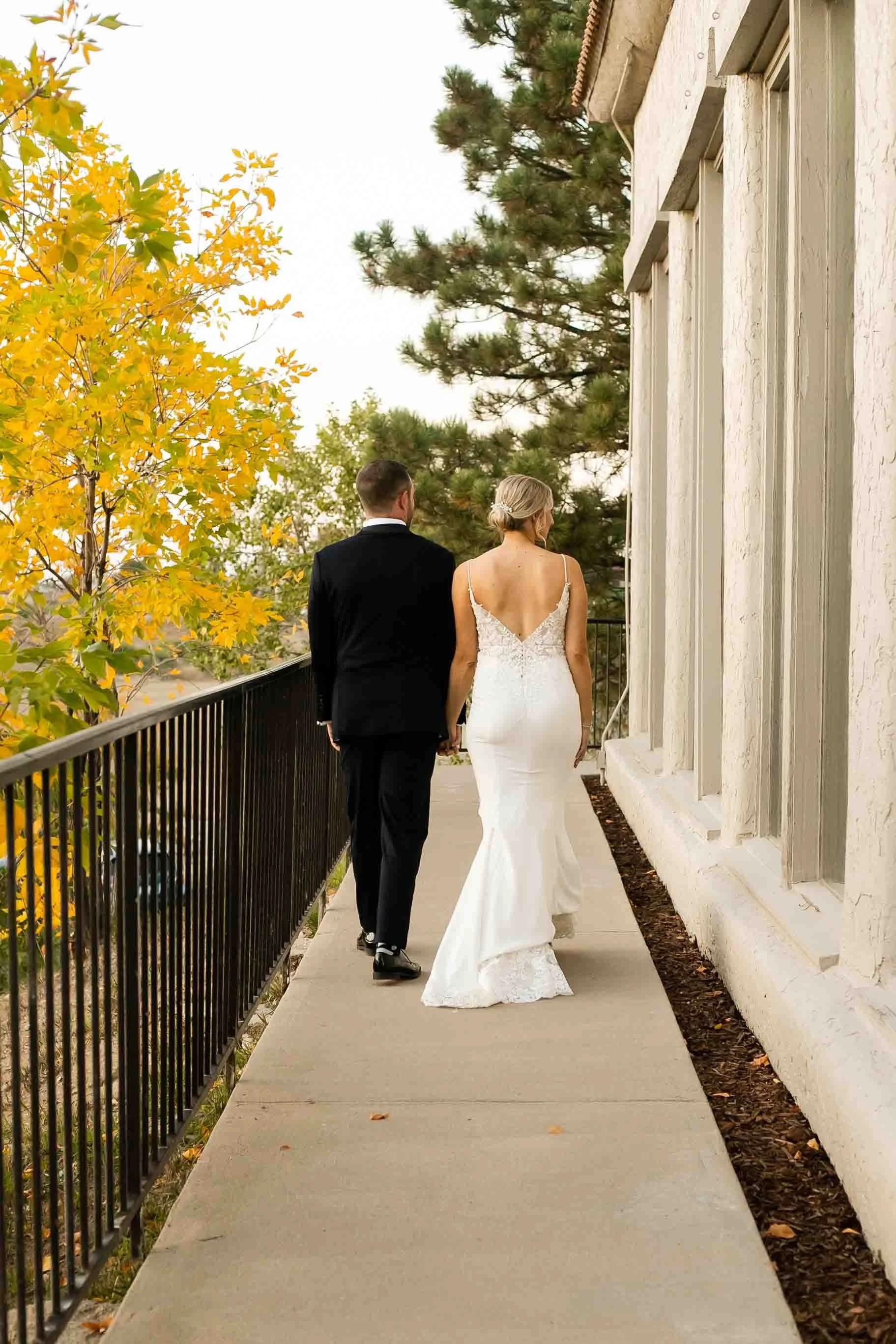 A bride and groom walking hand in hand on a balcony, with autumn trees in the background.