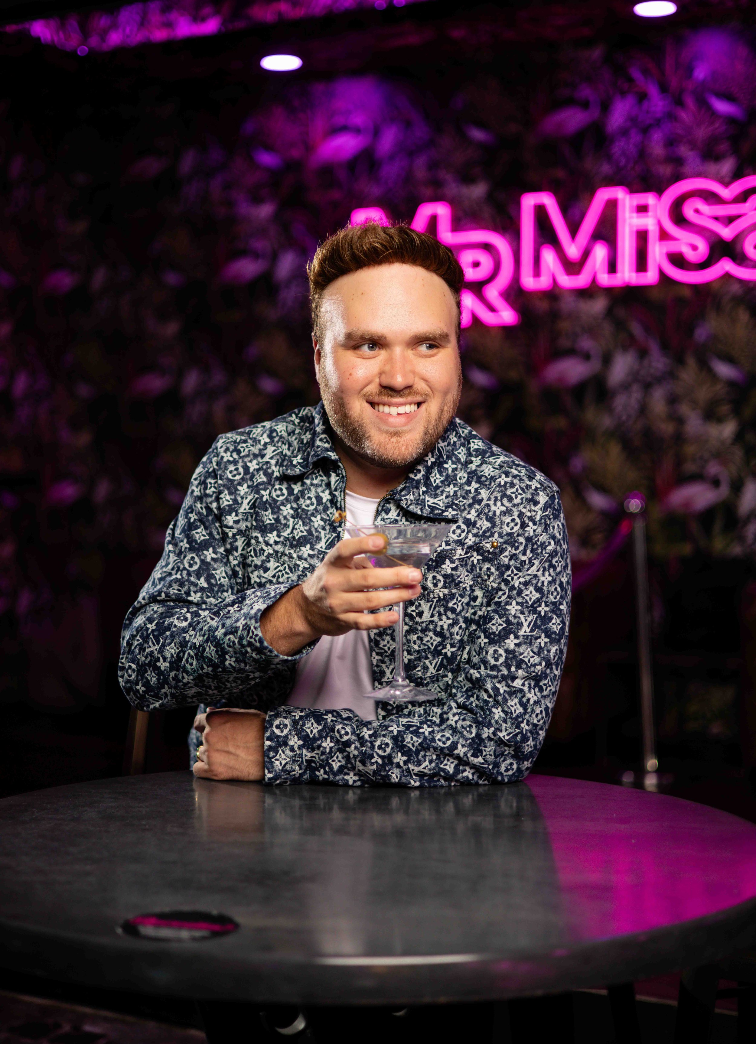 Person sitting in a bar holding a cocktail glass, with a neon sign display in the background.
