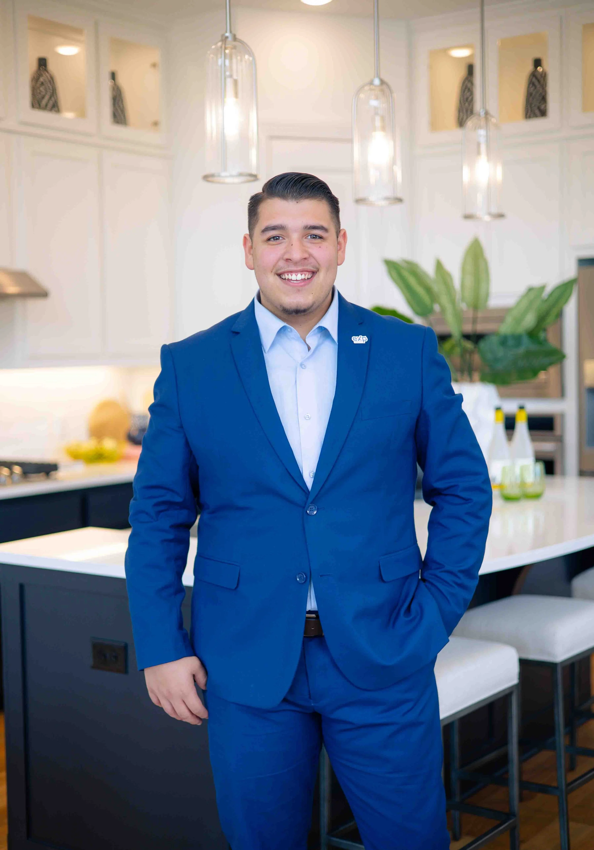 A young man wearing a blue suit and light blue shirt standing in a modern kitchen, smiling at the camera.
