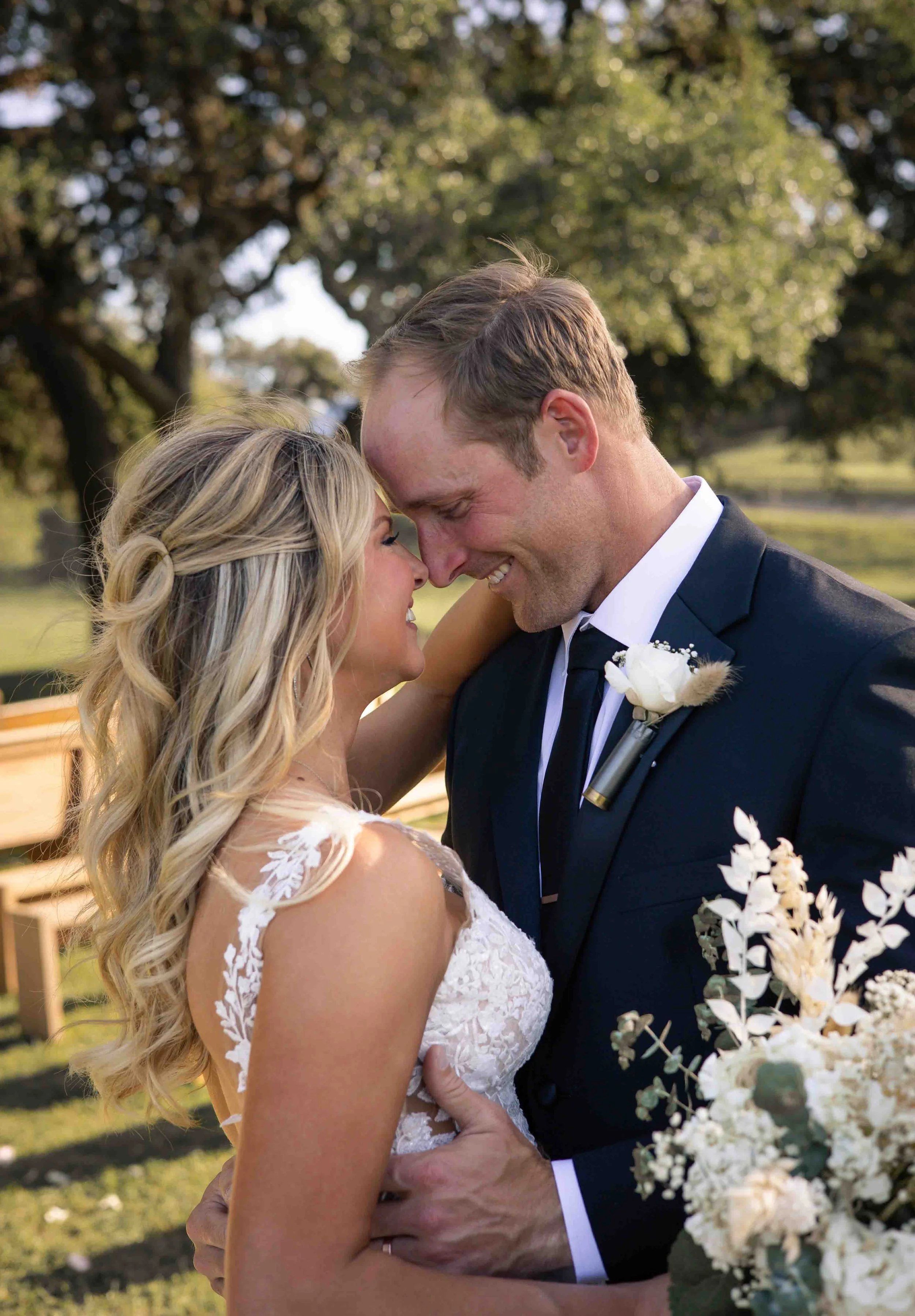 A bride and groom share an intimate moment outdoors on their wedding day, smiling and touching foreheads, with trees and sunlight in the background.