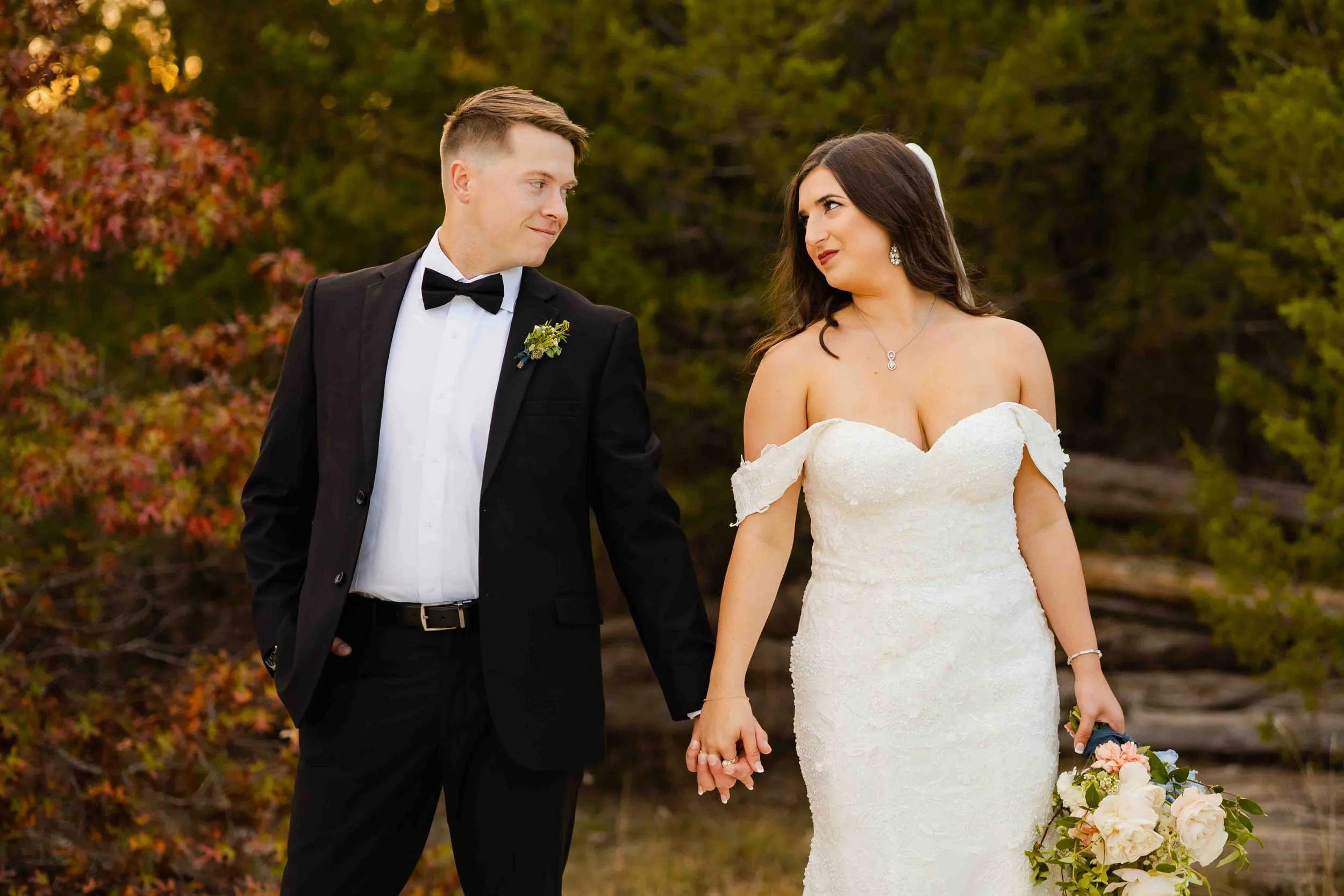 A bride and groom holding hands outdoors with autumn trees in the background. The bride wears a white, off-the-shoulder wedding gown and holds a bouquet of flowers. The groom wears a black tuxedo with a bow tie and boutonniere.