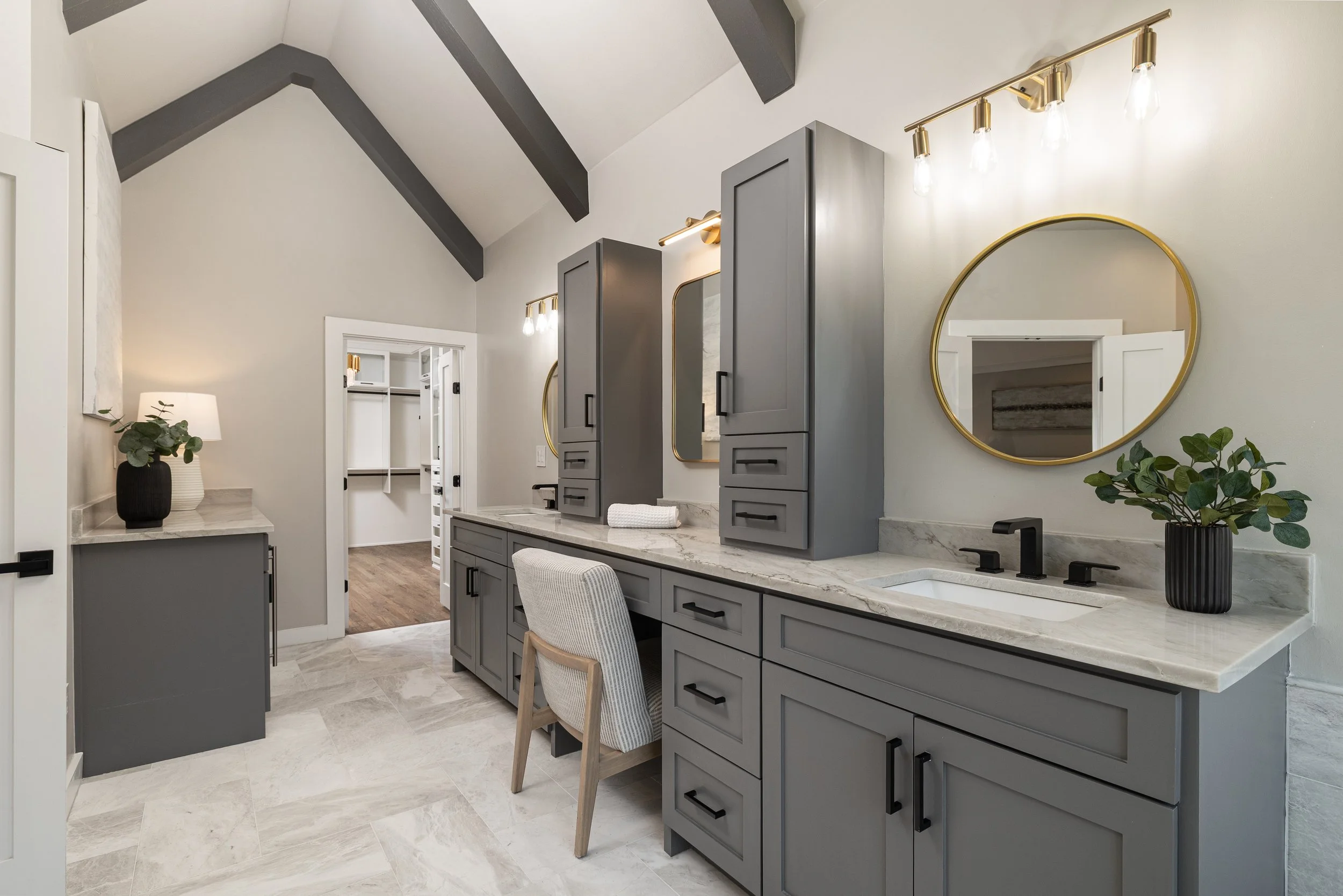Modern bathroom with gray cabinets, marble countertops, gold accents, and a round mirror; a chair is in front of the vanity, and there are potted plants on the counters.