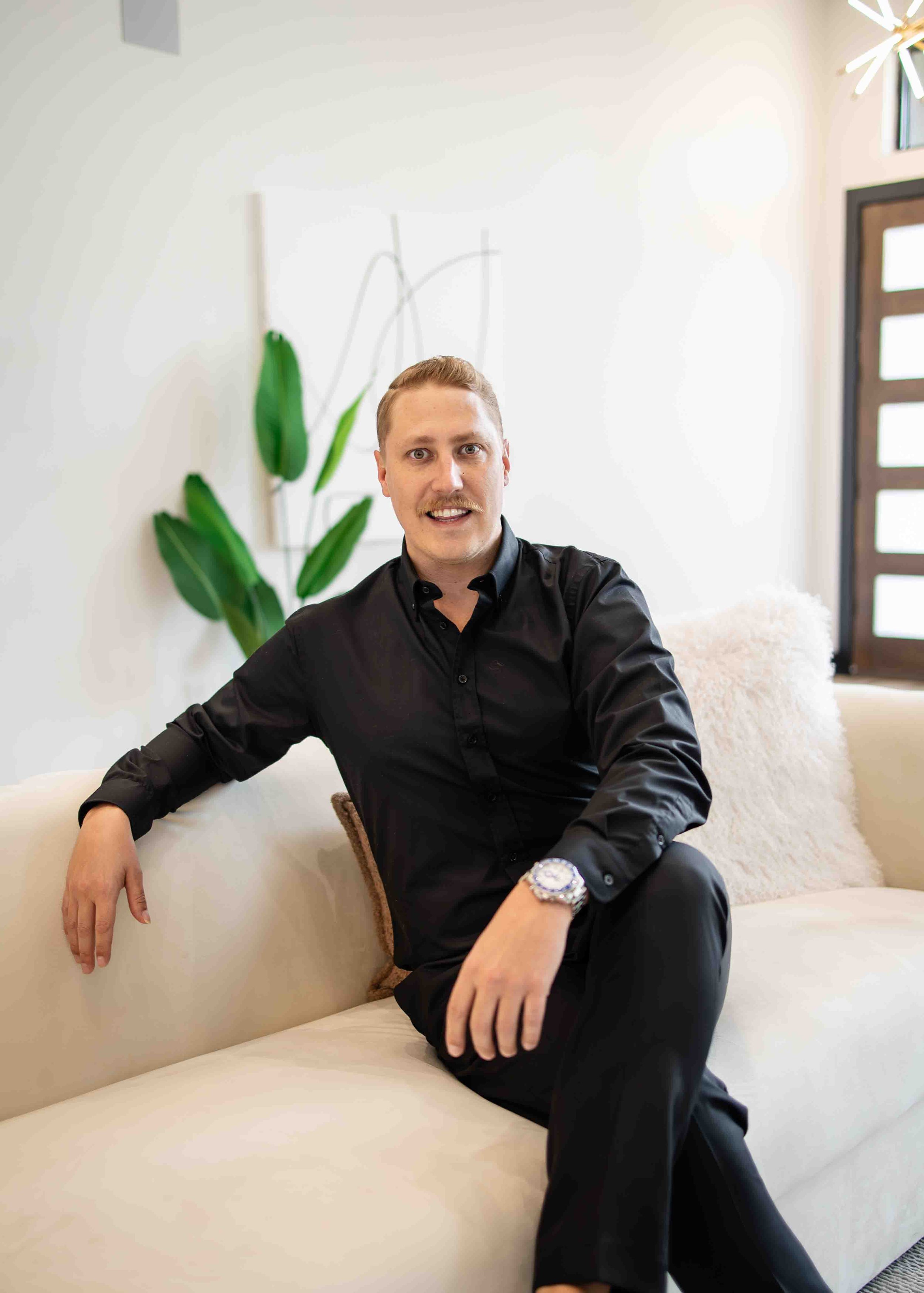 A man in a black shirt sitting on a white sofa in a modern living room, with plants and abstract artwork in the background.