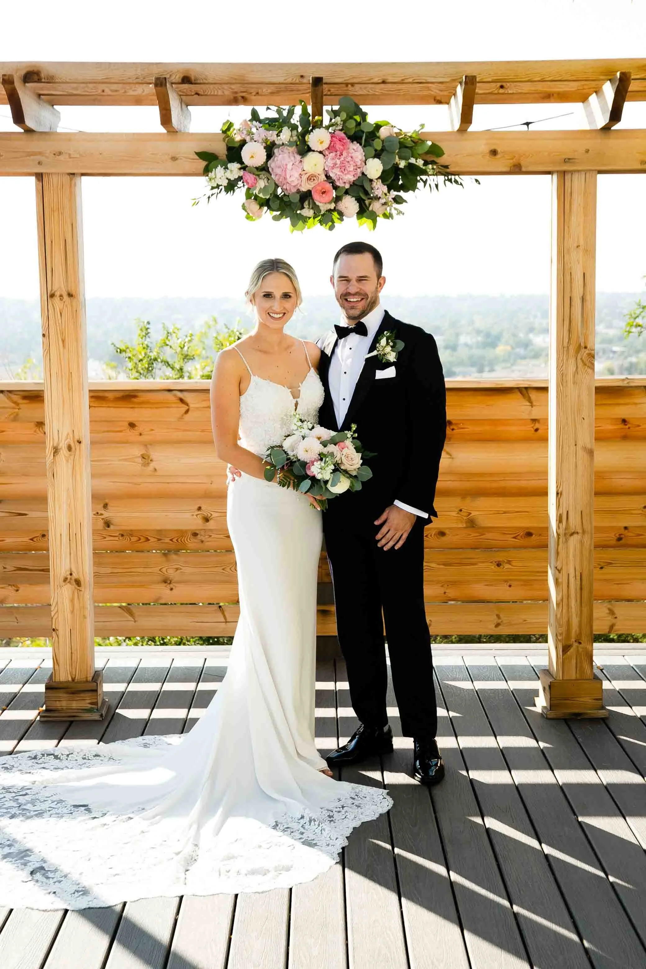 A bride and groom standing under a wooden arch with a floral arrangement of pink and white flowers hanging above them at outdoor wedding, with a scenic view in the background.