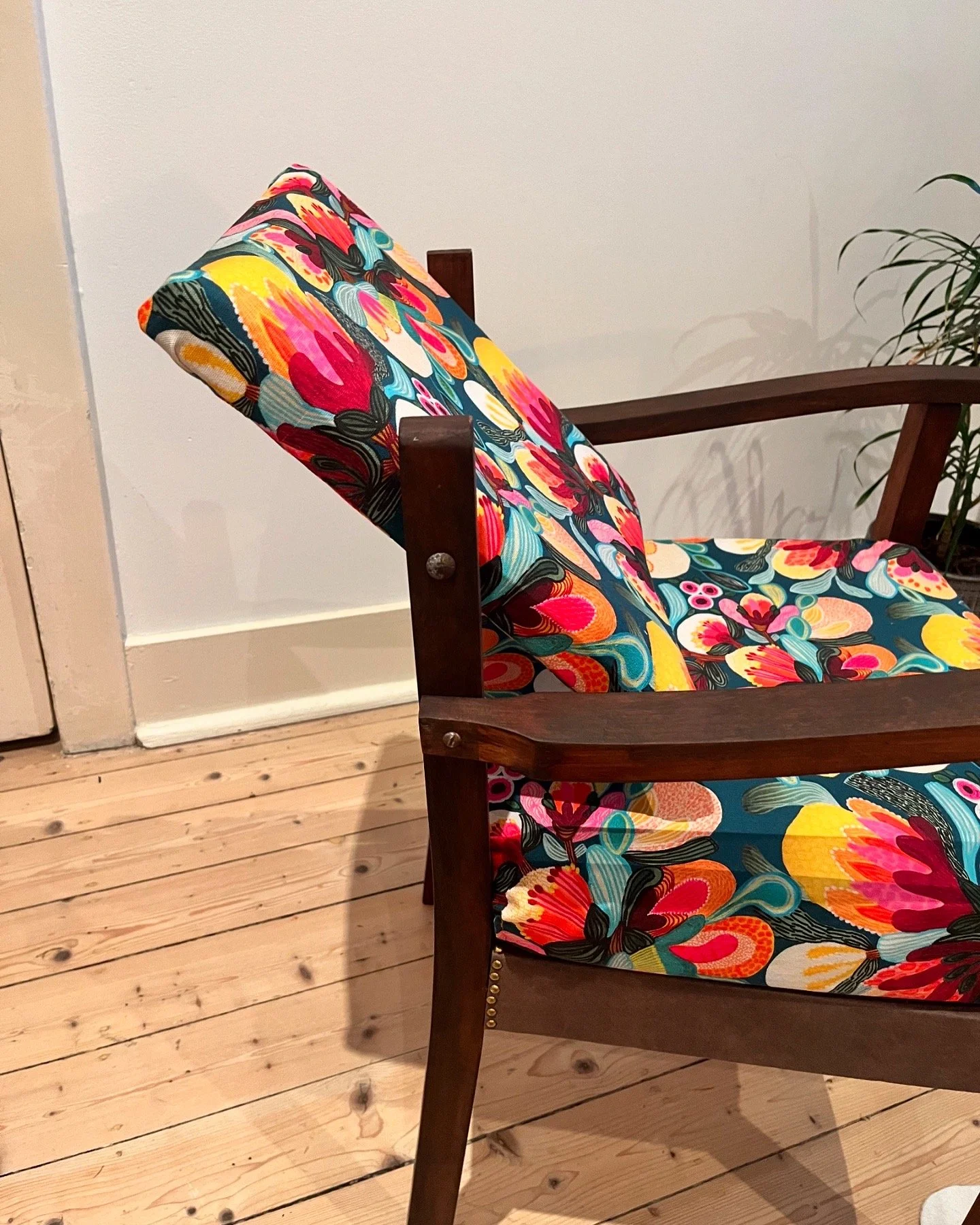 Mid-century modern chair with vibrant floral upholstery, wooden frame, on hardwood floor near a white wall and plant.