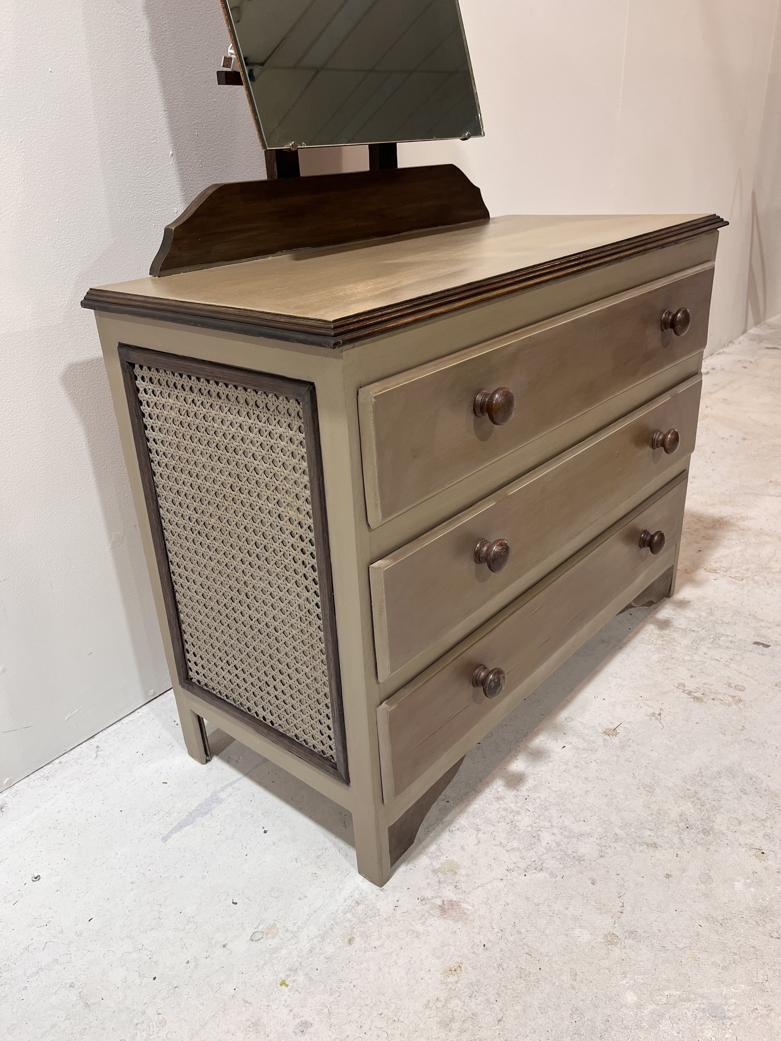 A vintage-style wooden dresser with three drawers and a woven cane side panel, topped with a tilting rectangular mirror on a wooden stand. The dresser has a light brown finish and round wooden knobs.