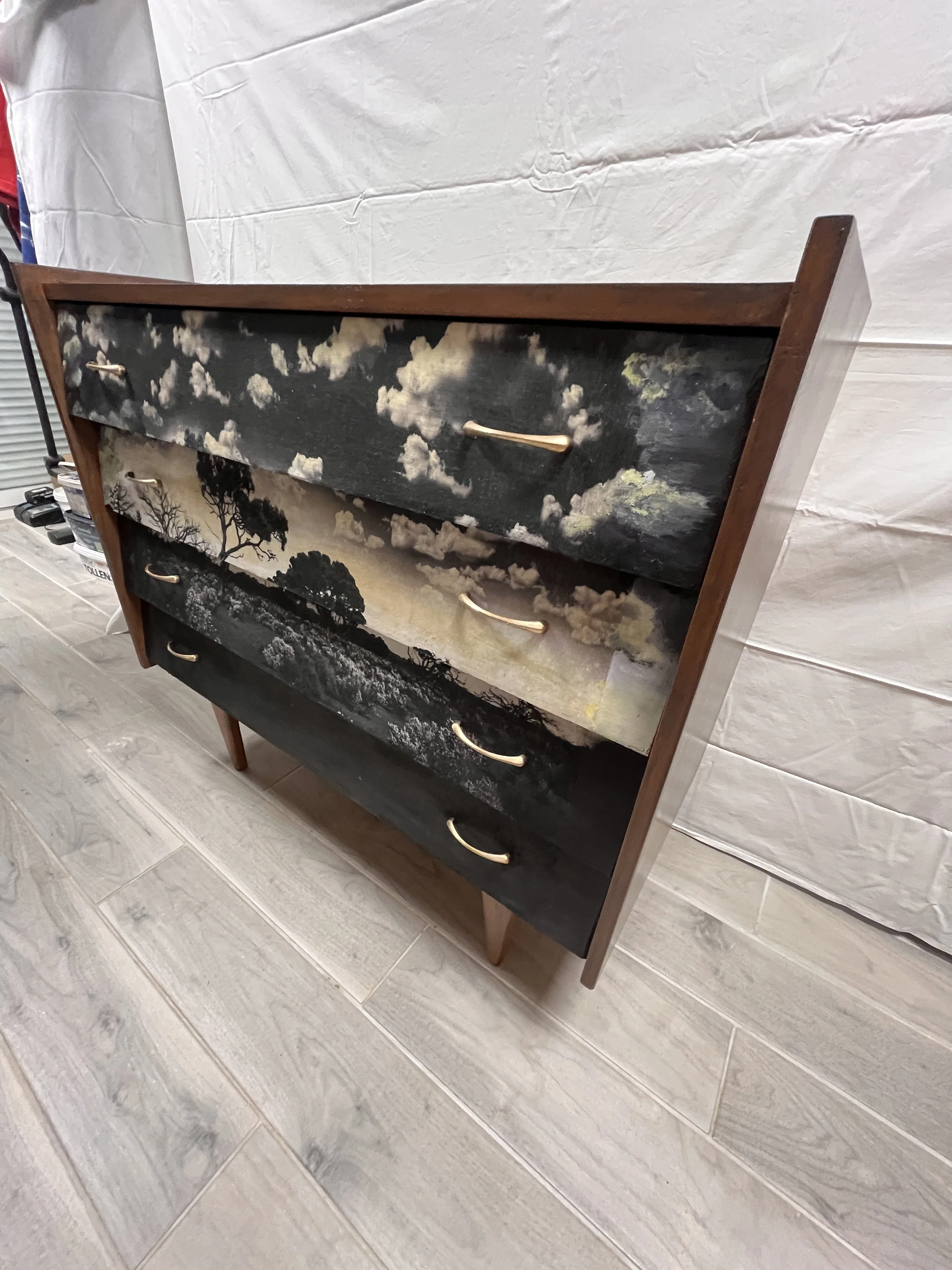 A wooden dresser with three drawers, each featuring landscape paintings of clouds and trees in black and white.