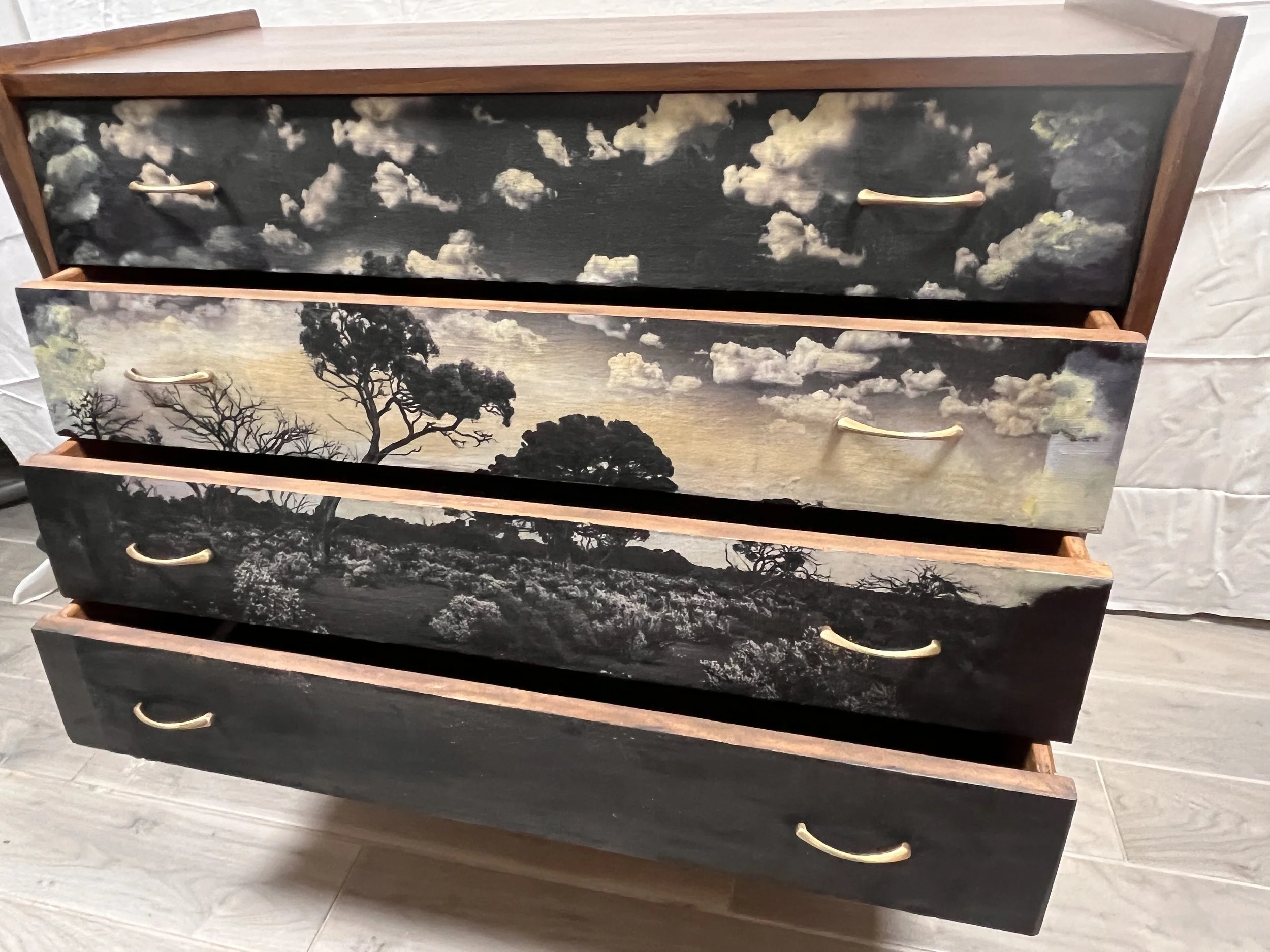 A four-drawer wooden dresser with painted landscape scenes on the drawer fronts, featuring trees, clouds, and a sky in black, gray, and beige tones.