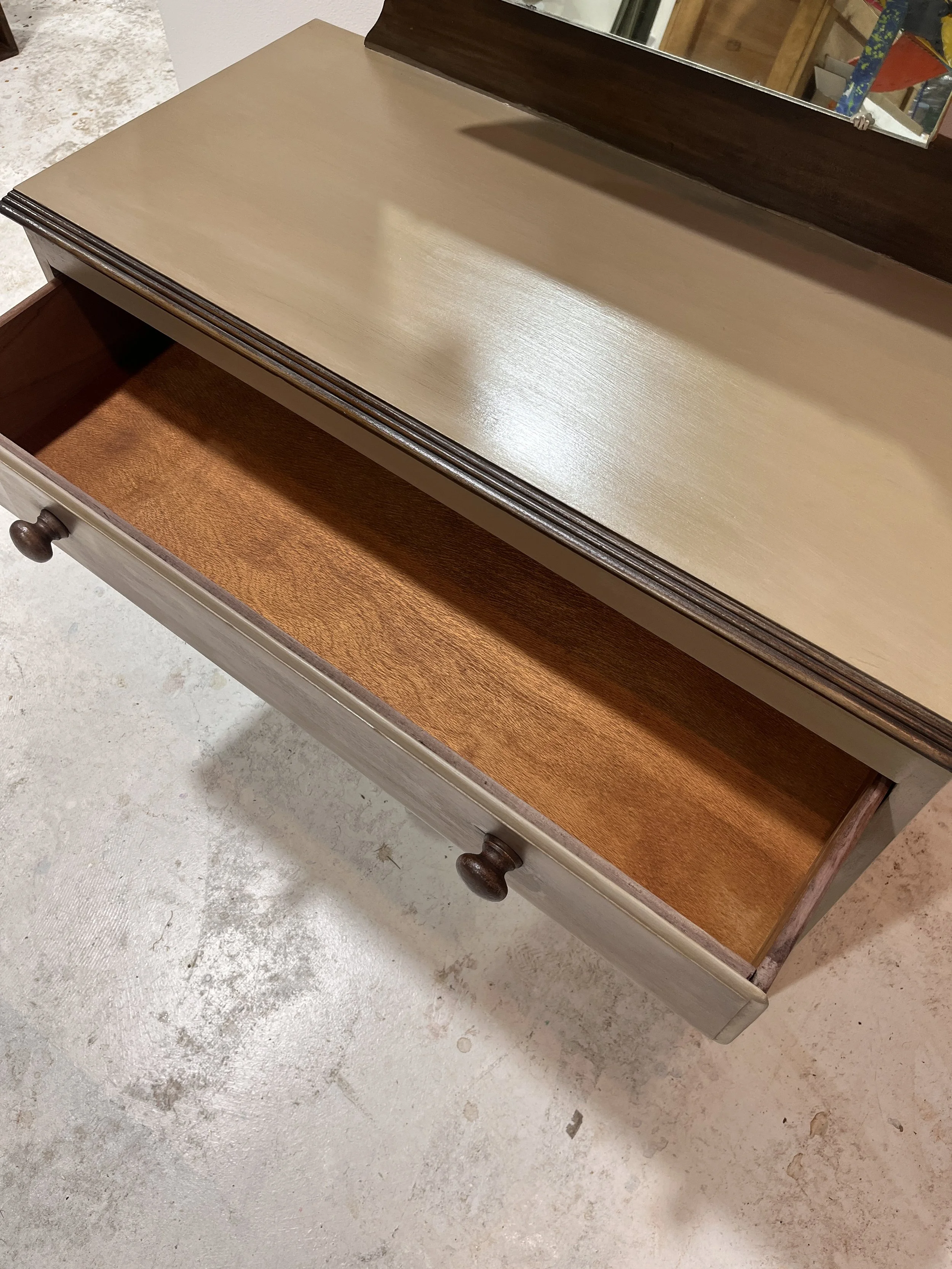 Open wooden drawer in a beige dresser with a mirror attached, displayed on a concrete floor.