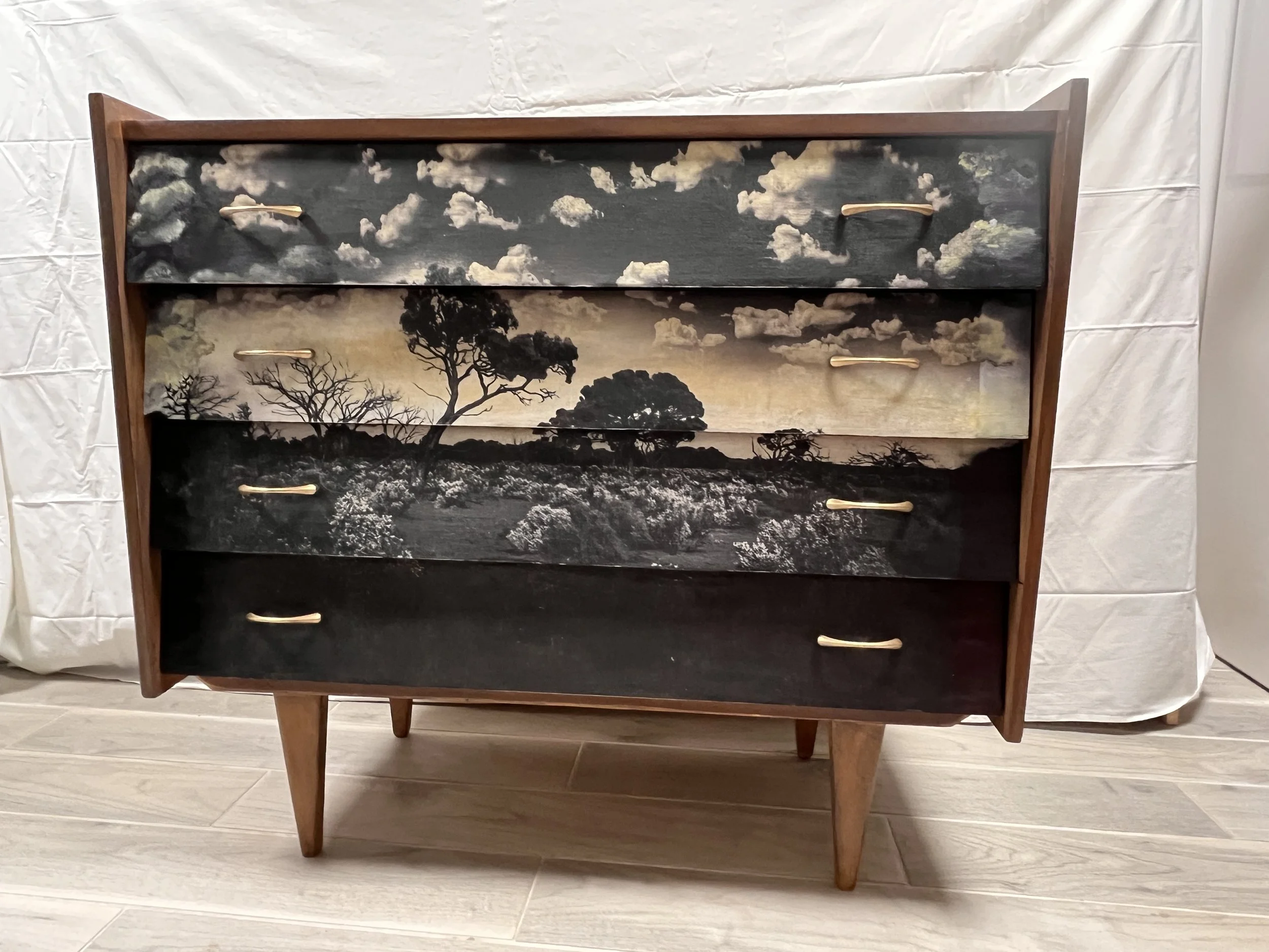 Mid-century modern wooden dresser with five drawers, each featuring a landscape scene with trees and sky painted on the fronts. The dresser is on tapered legs and is placed on a light-colored wood floor against a white backdrop.