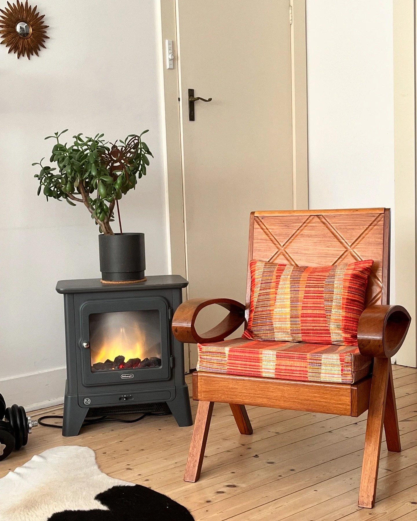 Cozy corner with a wooden chair featuring red and orange striped cushions, an electric fireplace with a potted plant on top, and a decorative wall mirror on a light-colored wall.