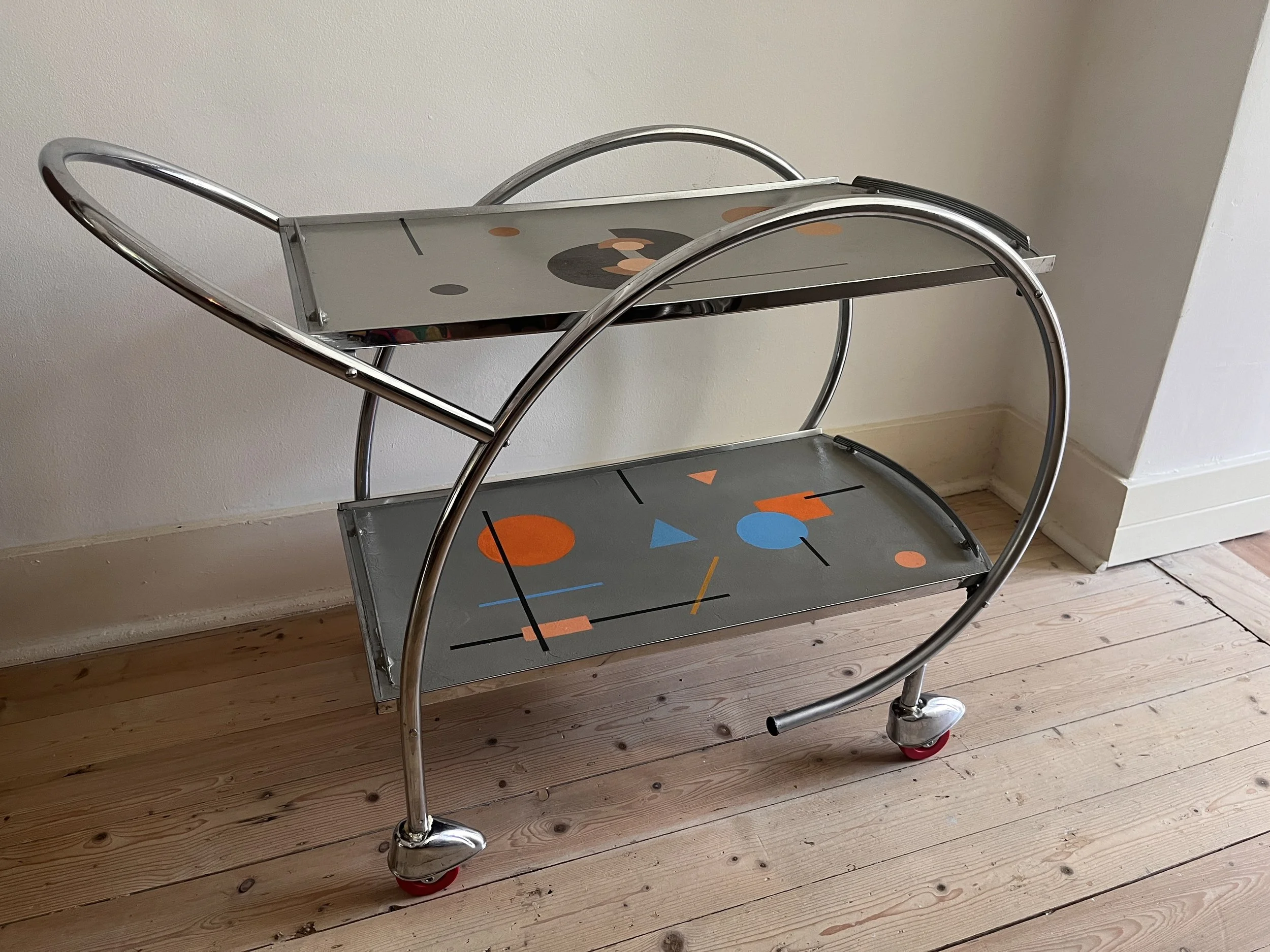 Retro tea trolley with two glass shelves featuring geometric patterns, chrome frame, and wheels, on a wooden floor.