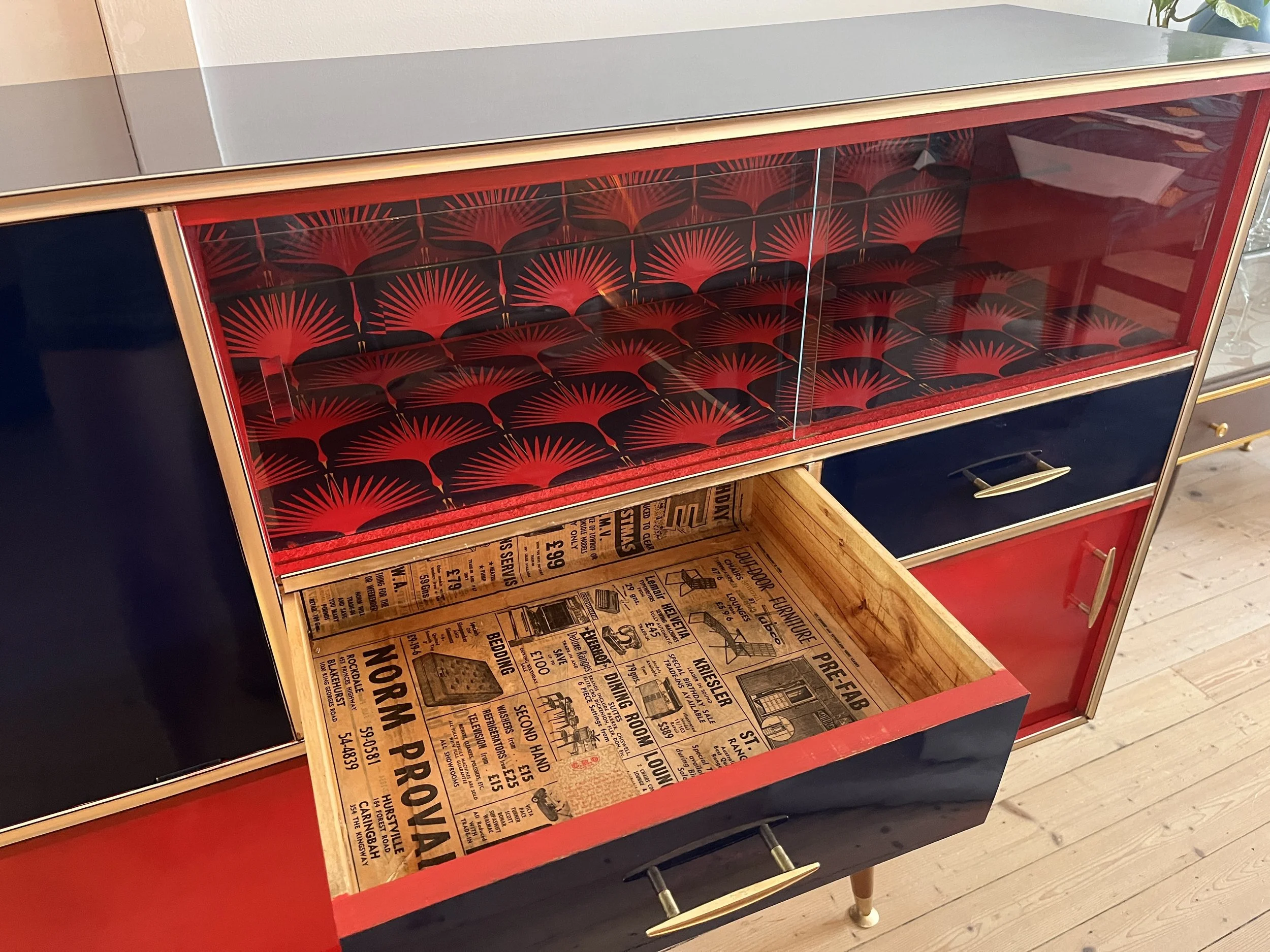 Vintage cabinet with red and navy design, featuring a drawer lined with newspaper print and a glass-fronted section with a red decorative pattern.