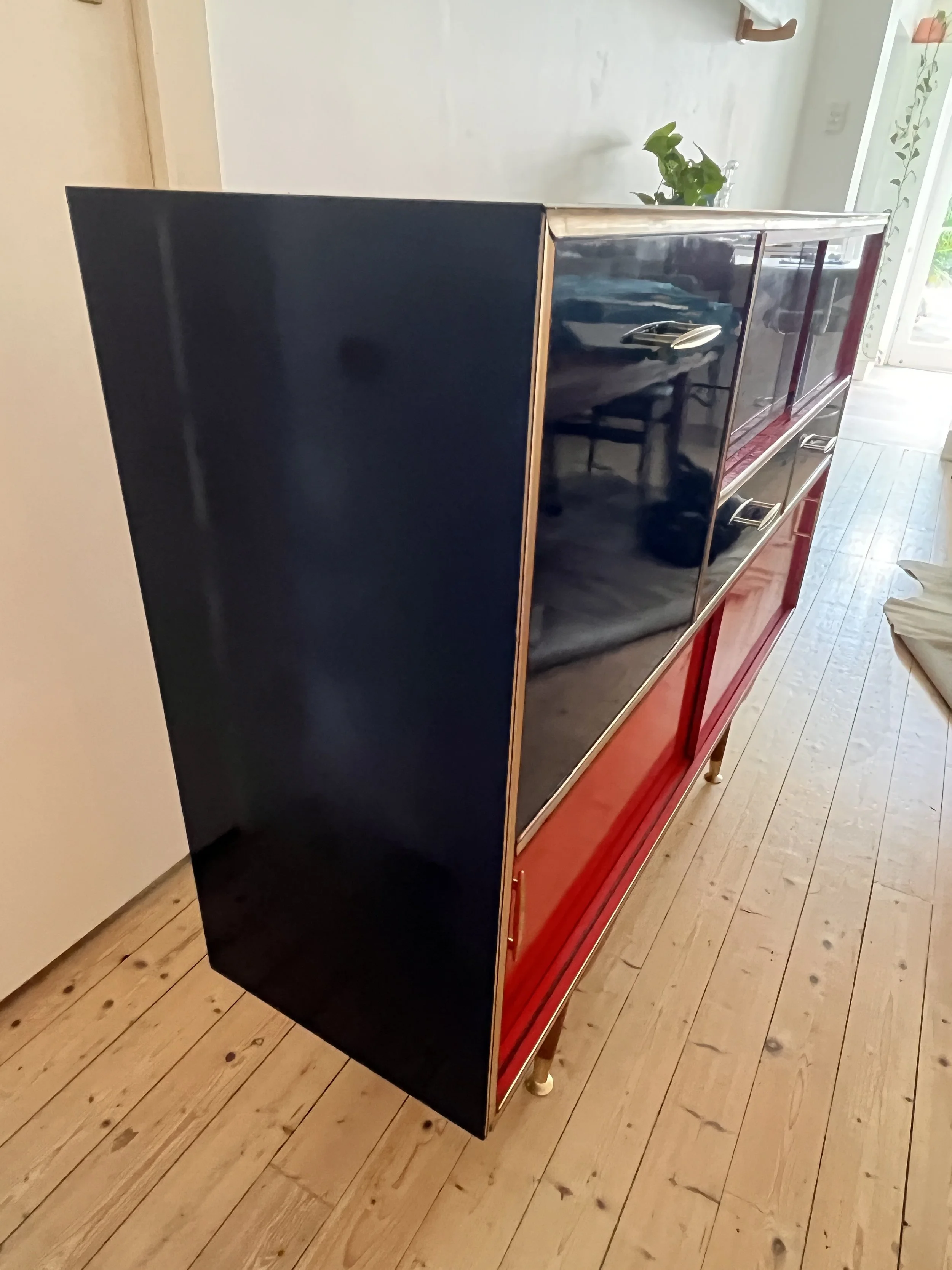 A modern sideboard with a glossy finish, featuring dark blue and red panels, gold handles, and legs, situated on a wooden floor.