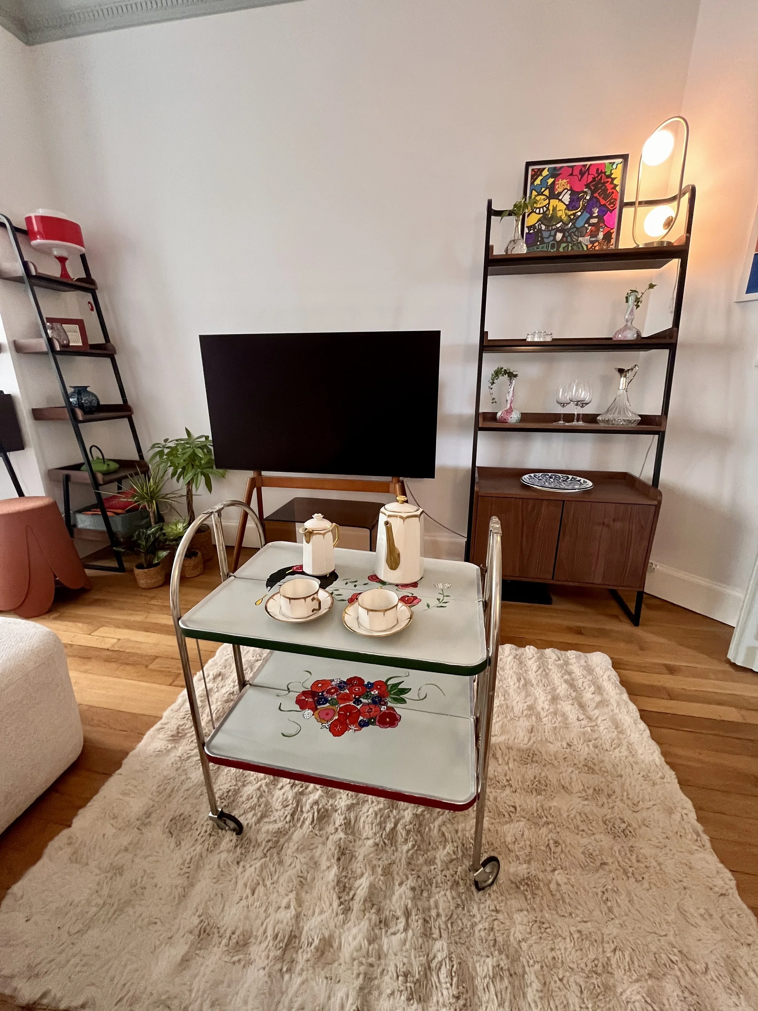 Living room with a wall-mounted flat-screen TV, wooden floor, beige shaggy rug, vintage rolling tea cart with floral design, teapots, cups, a small black table, potted plants, bookshelves with decorative items, and wall art.