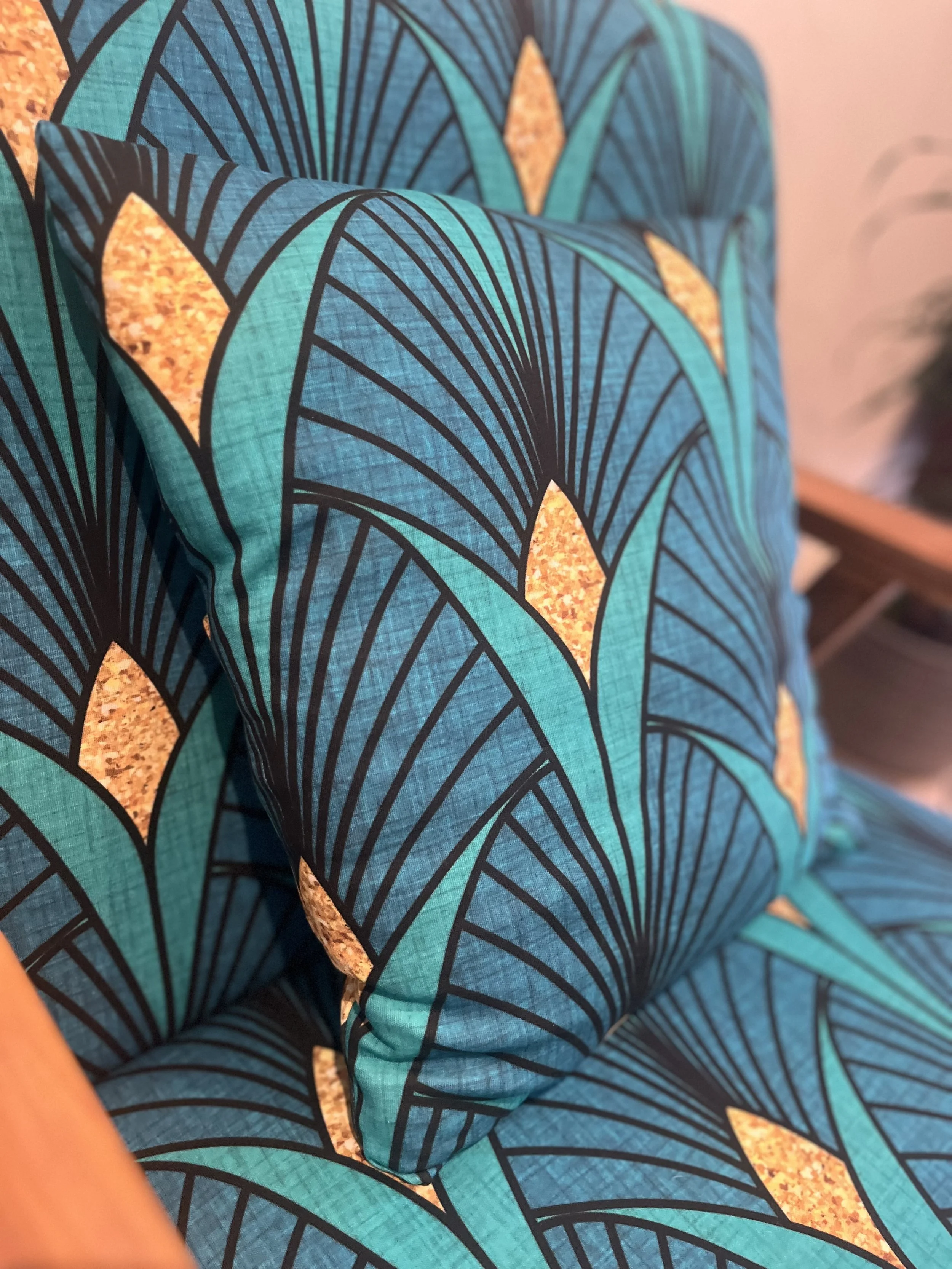 A close-up view of a decorative pillow on a chair, featuring a teal and black geometric pattern with gold leaf-like shapes.