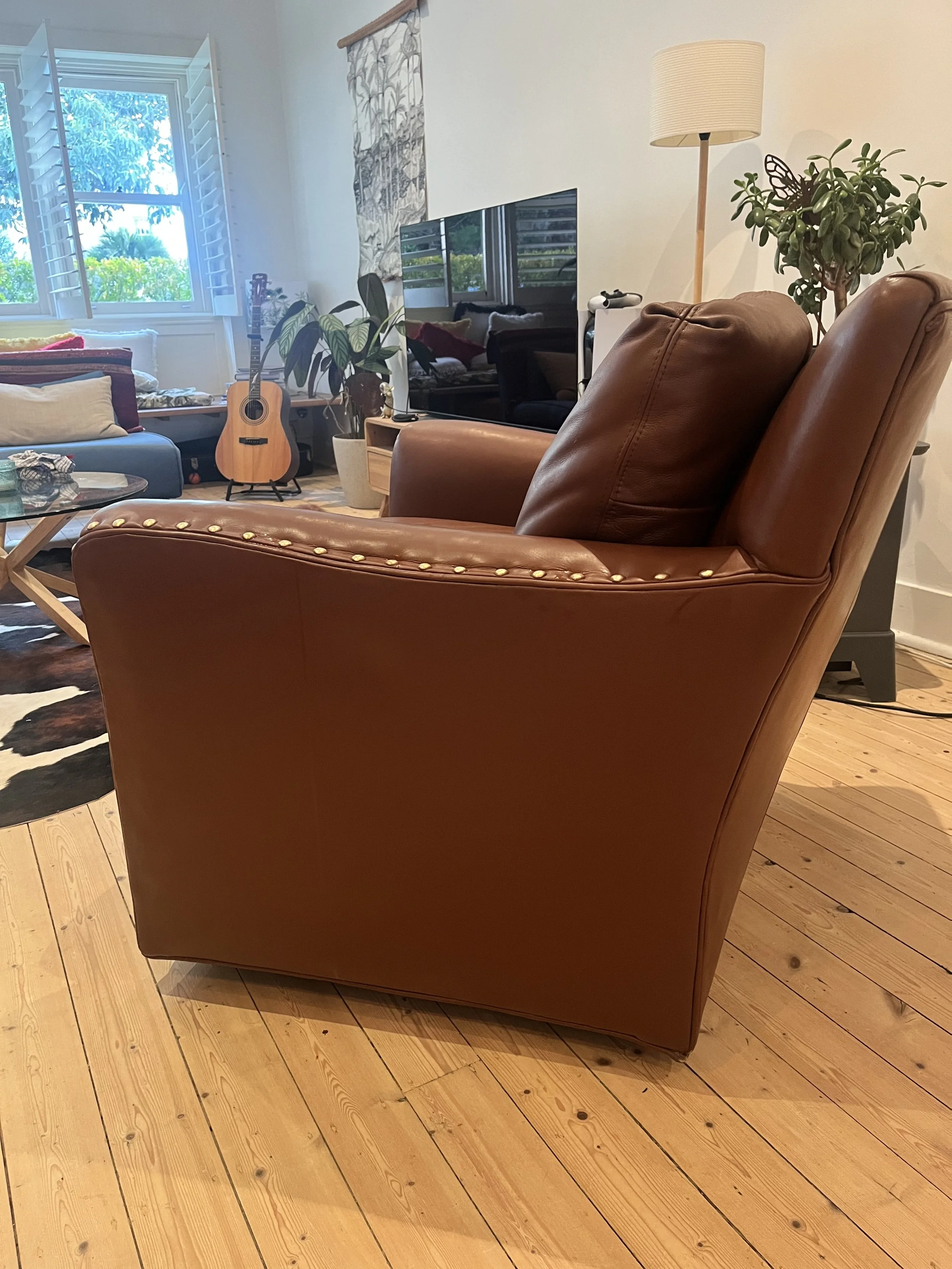 Living room with brown leather armchair, wooden floor, TV, guitar, couch, potted plants, and wall decor.