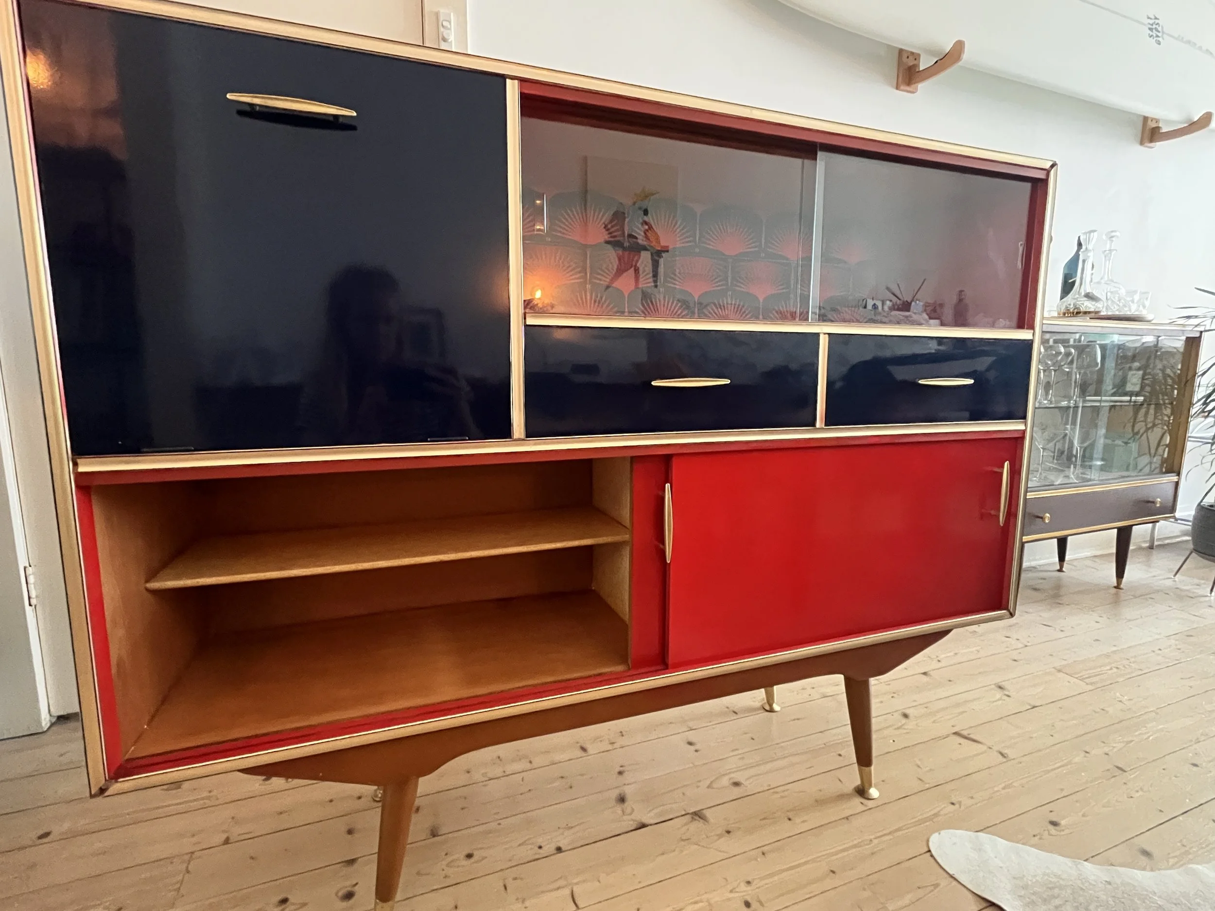 Mid-century modern cabinet with glass doors and shelves, featuring red, black, and wooden finish, standing on tapered legs, decorated with glassware and a background wallpaper.