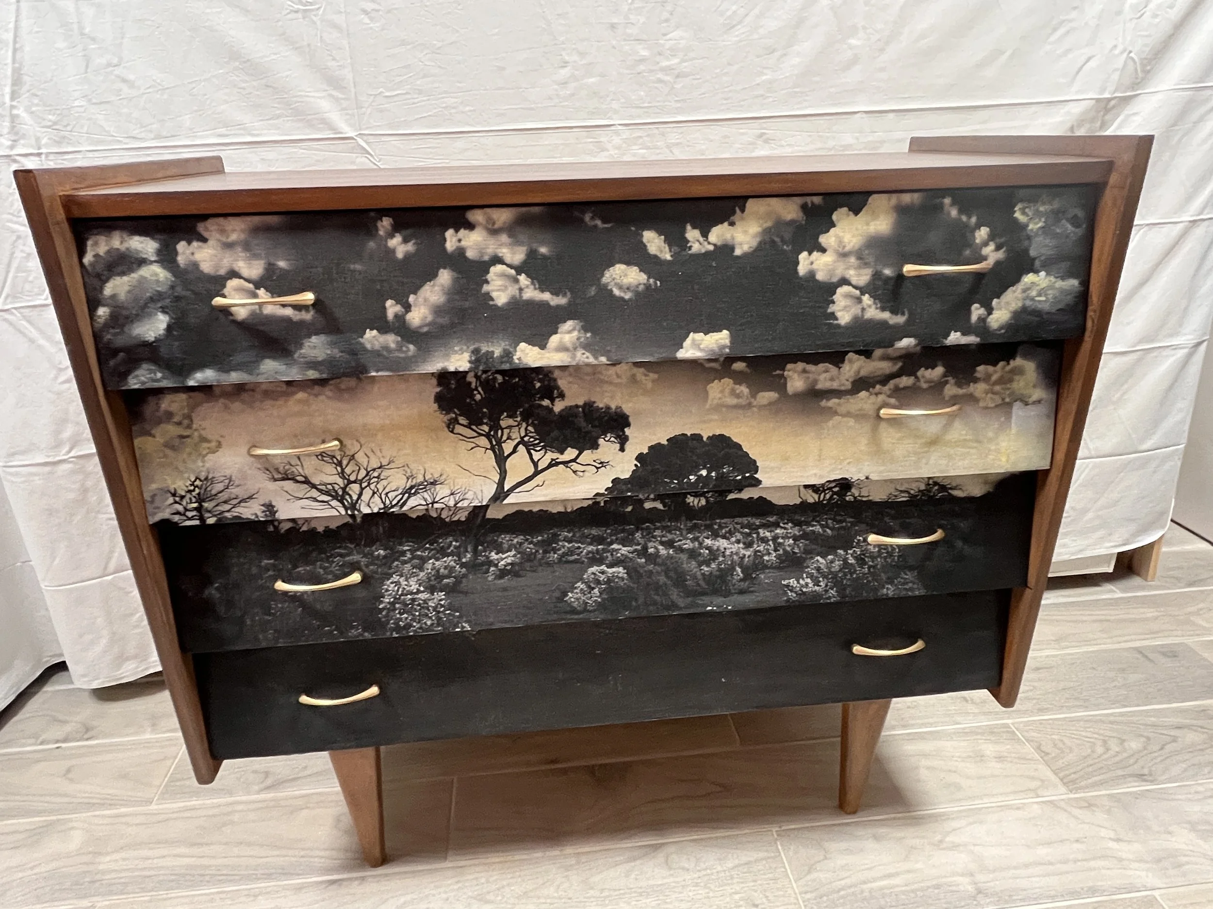 A wooden dresser with four drawers painted with a landscape scene of trees and clouds, featuring black, white, and beige colors.