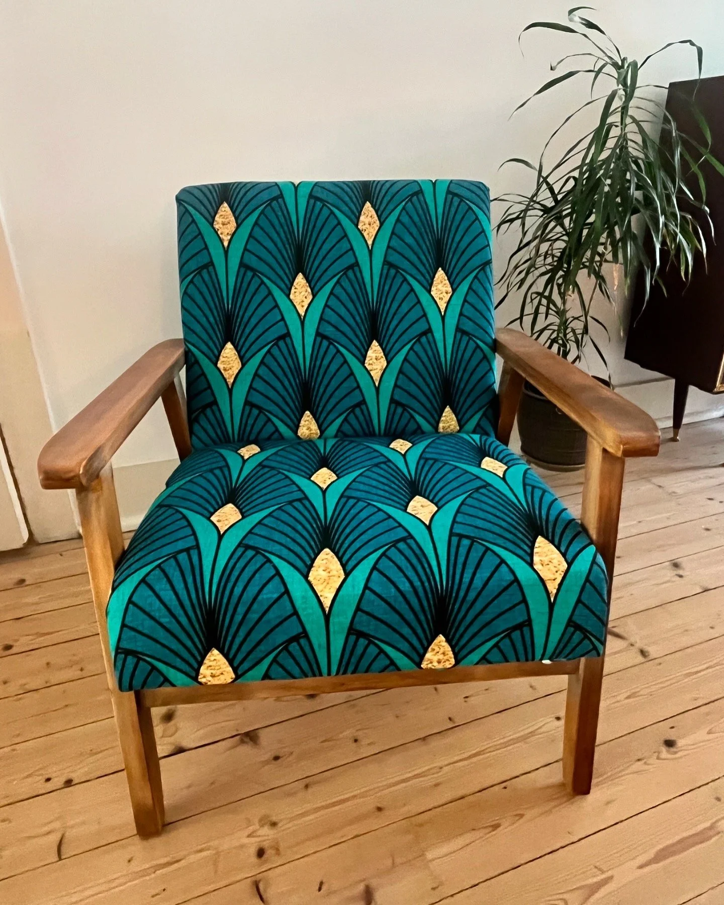 Mid-century modern chair with colorful geometric print upholstery and wooden armrests on a wooden floor, with a potted plant in the background.