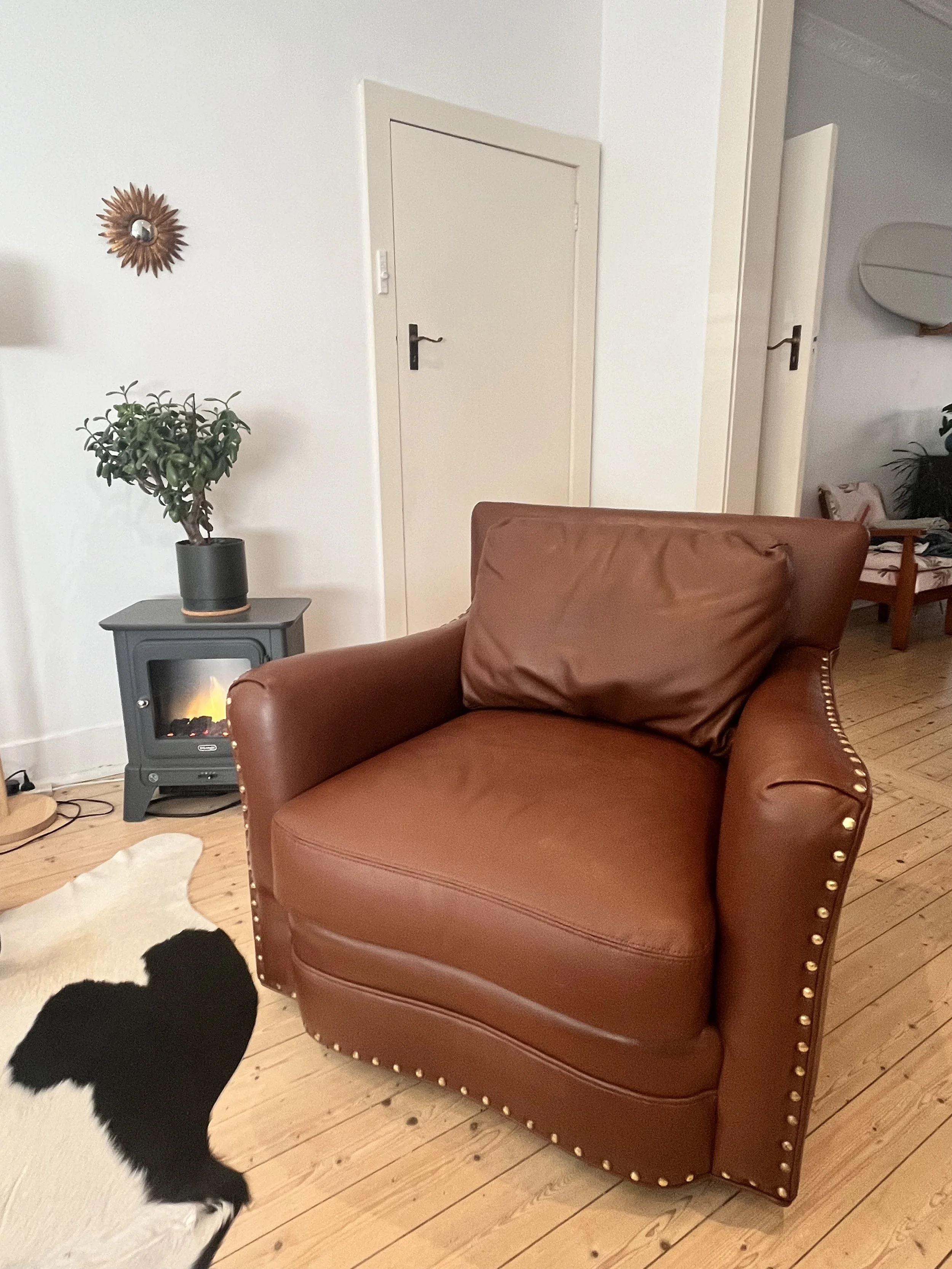 Brown leather armchair with brass studs, hardwood floor, cowhide rug, small stove with electric fire, potted plant, round sunburst wall mirror in a cozy room.